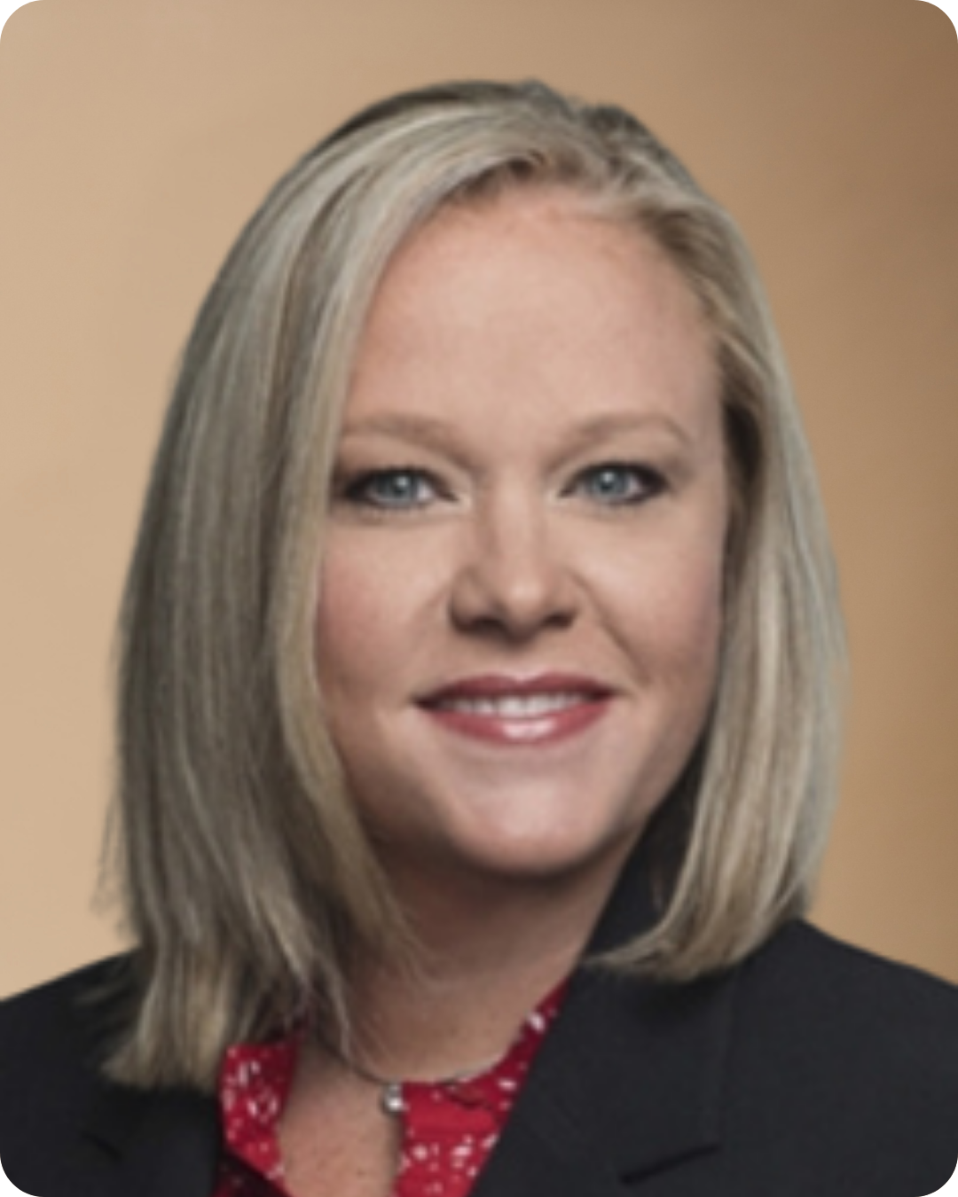 A portrait photo of a woman with shoulder-length blonde hair, blue eyes, wearing a black blazer and a red patterned blouse, smiling against a plain beige background.
