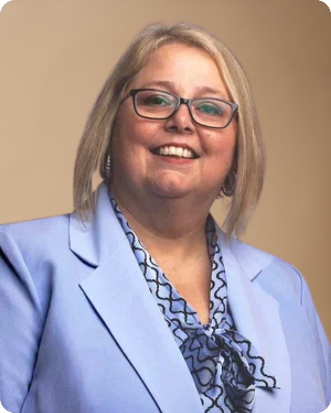 A woman with blonde hair, glasses, and earrings wearing a light blue blazer and patterned blouse, smiling against a plain background.