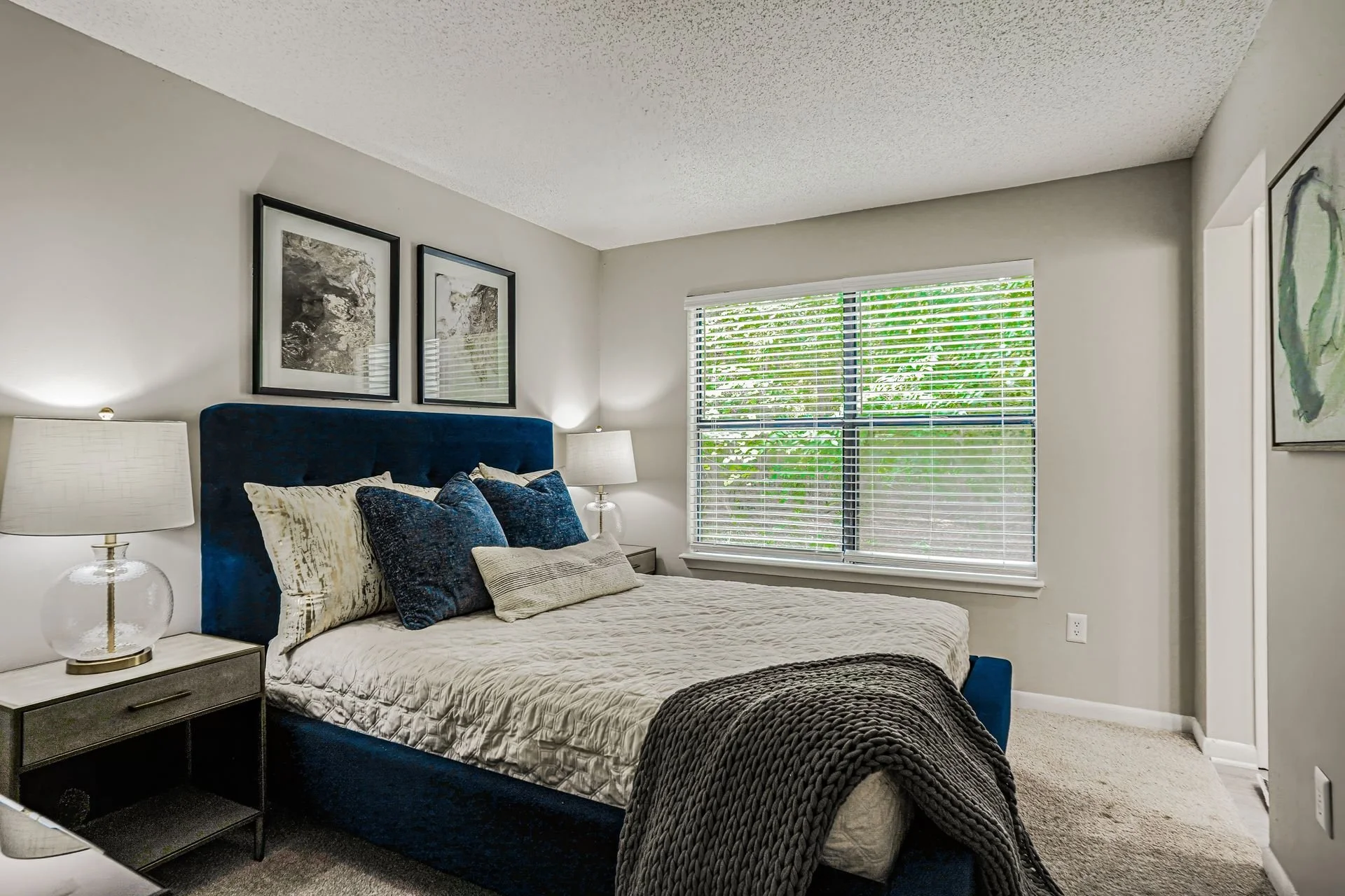 Bedroom with white walls, large window with blinds, blue upholstered bed, pillows, bedside table with lamp, wall art, and carpeted floor.