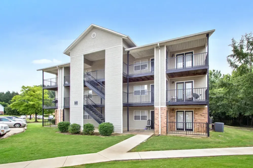 Multi-story apartment building with outdoor staircases, balconies with black railings, and a grassy lawn area.