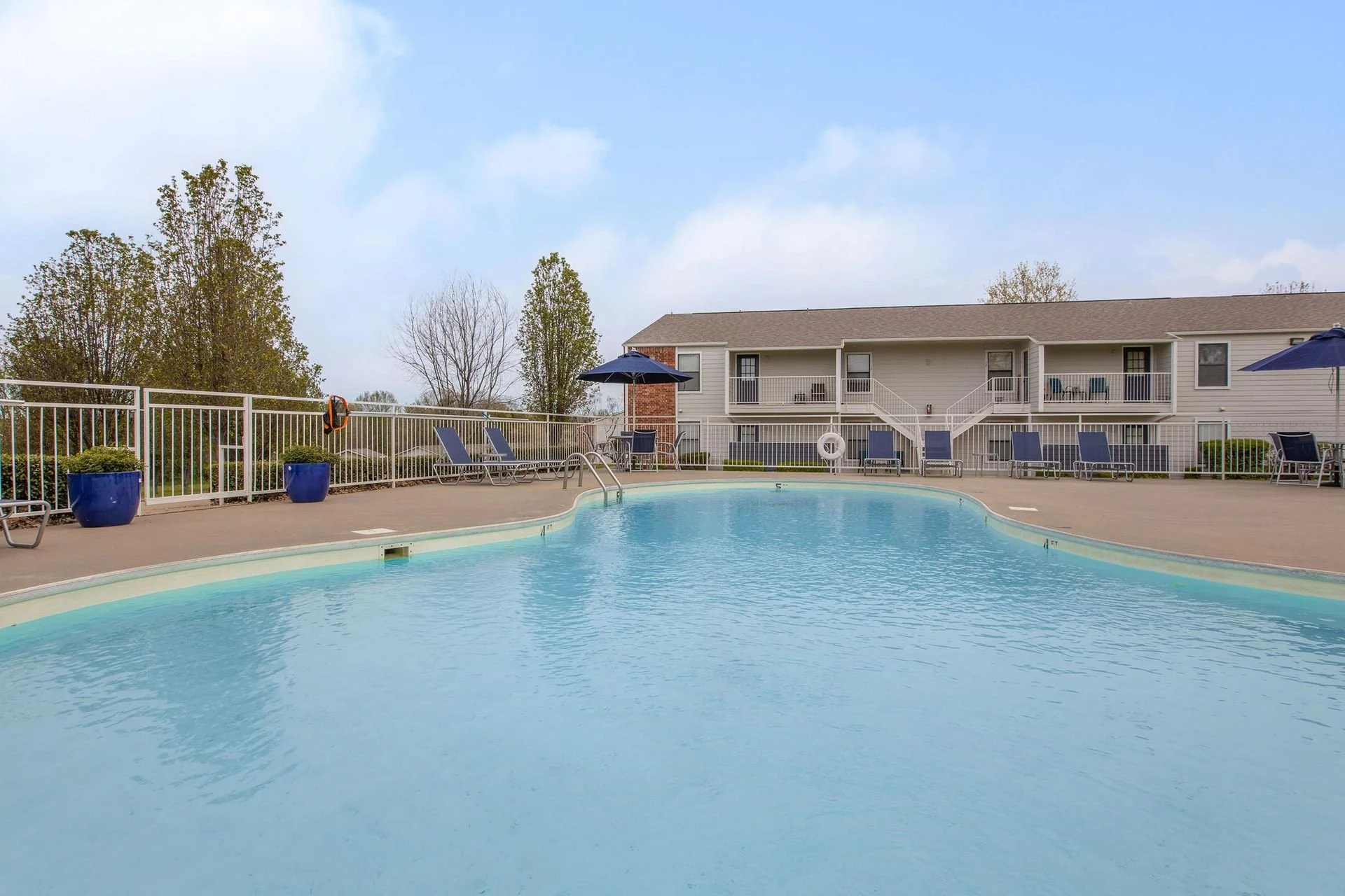 Empty outdoor swimming pool surrounded by lounge chairs, umbrellas, and potted plants in a residential complex with apartment buildings and trees in the background.