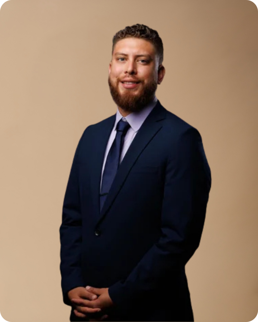 A man with a beard and short hair wearing a dark suit, white shirt, and tie, standing against a plain beige background.
