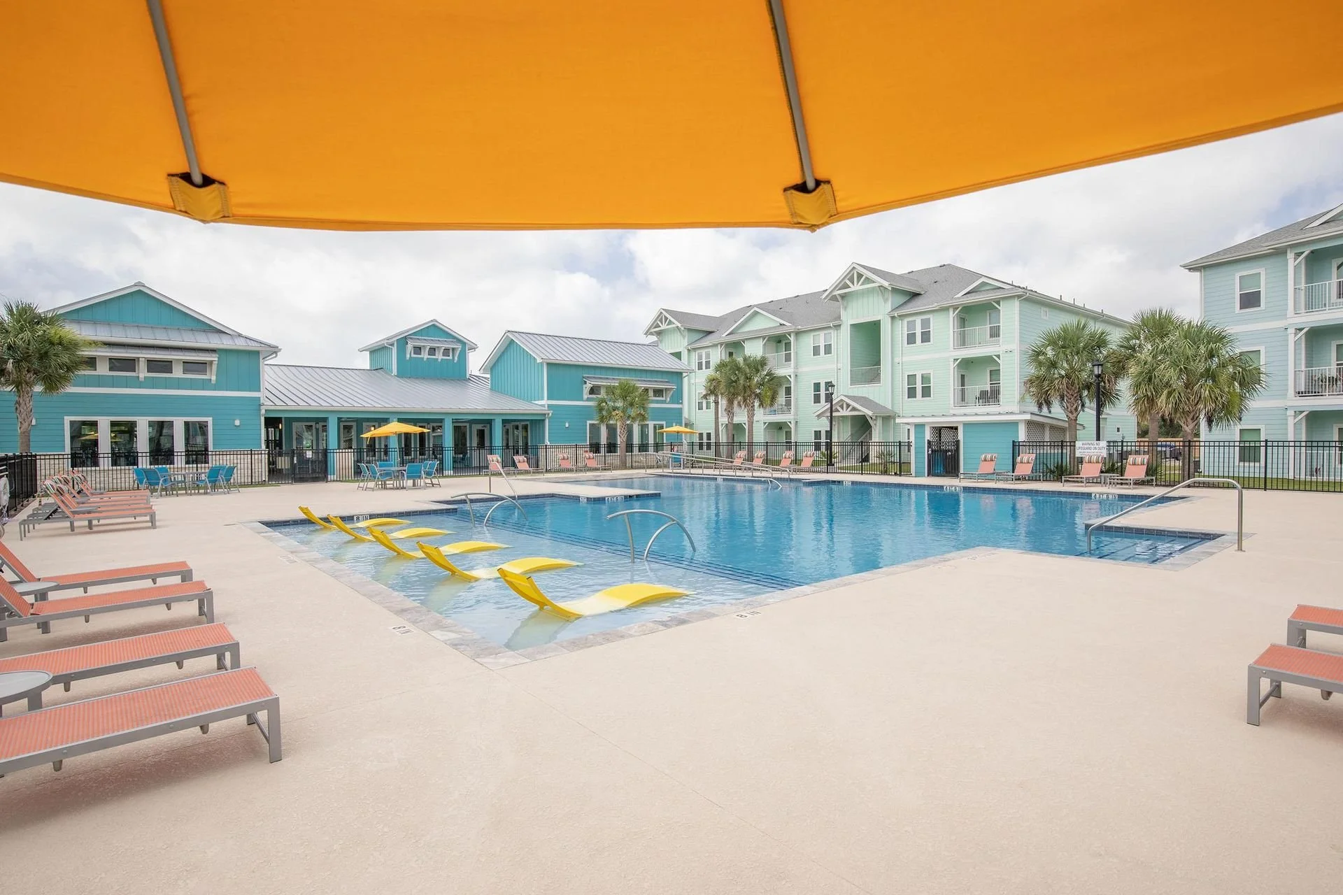 View of a swimming pool with yellow slides, lounge chairs, and umbrellas at a colorful residential complex with pastel blue and green buildings and palm trees.