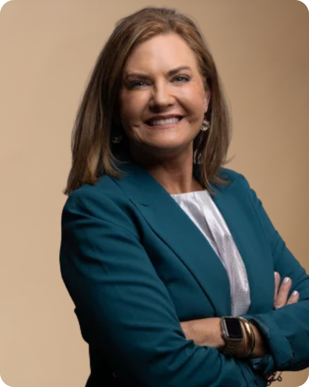 A woman with light skin, brown hair, and a big smile, wearing a teal blazer, white blouse, earrings, and a smartwatch, standing with crossed arms against a beige background.