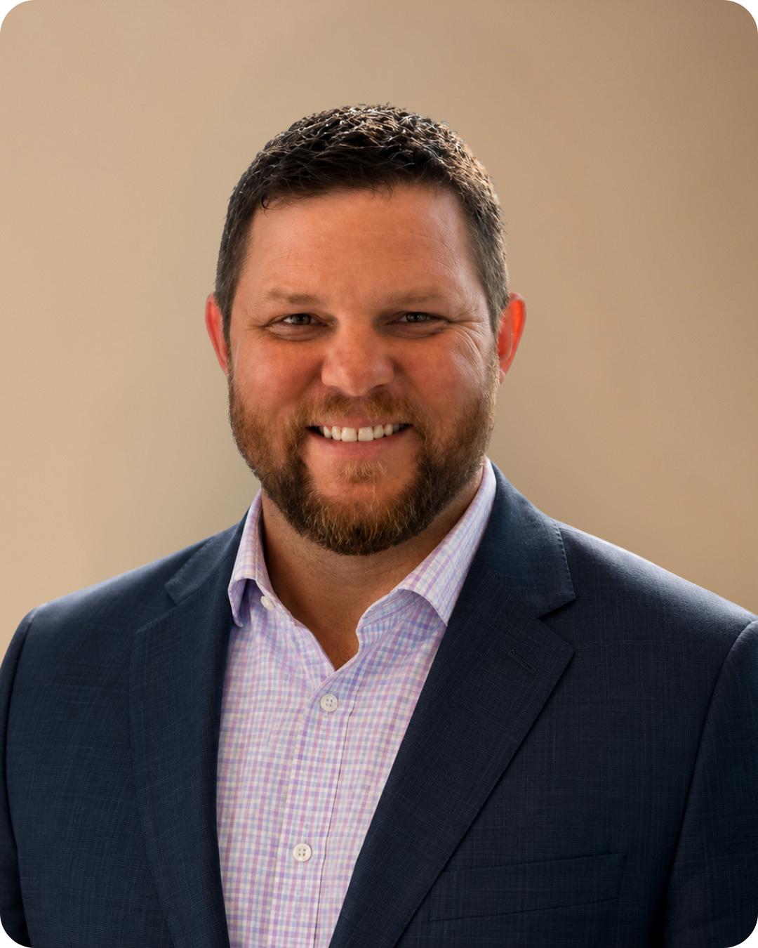 Headshot of a smiling man with a beard and short dark hair, wearing a dark blue suit jacket and a light pink plaid dress shirt, on a beige background.