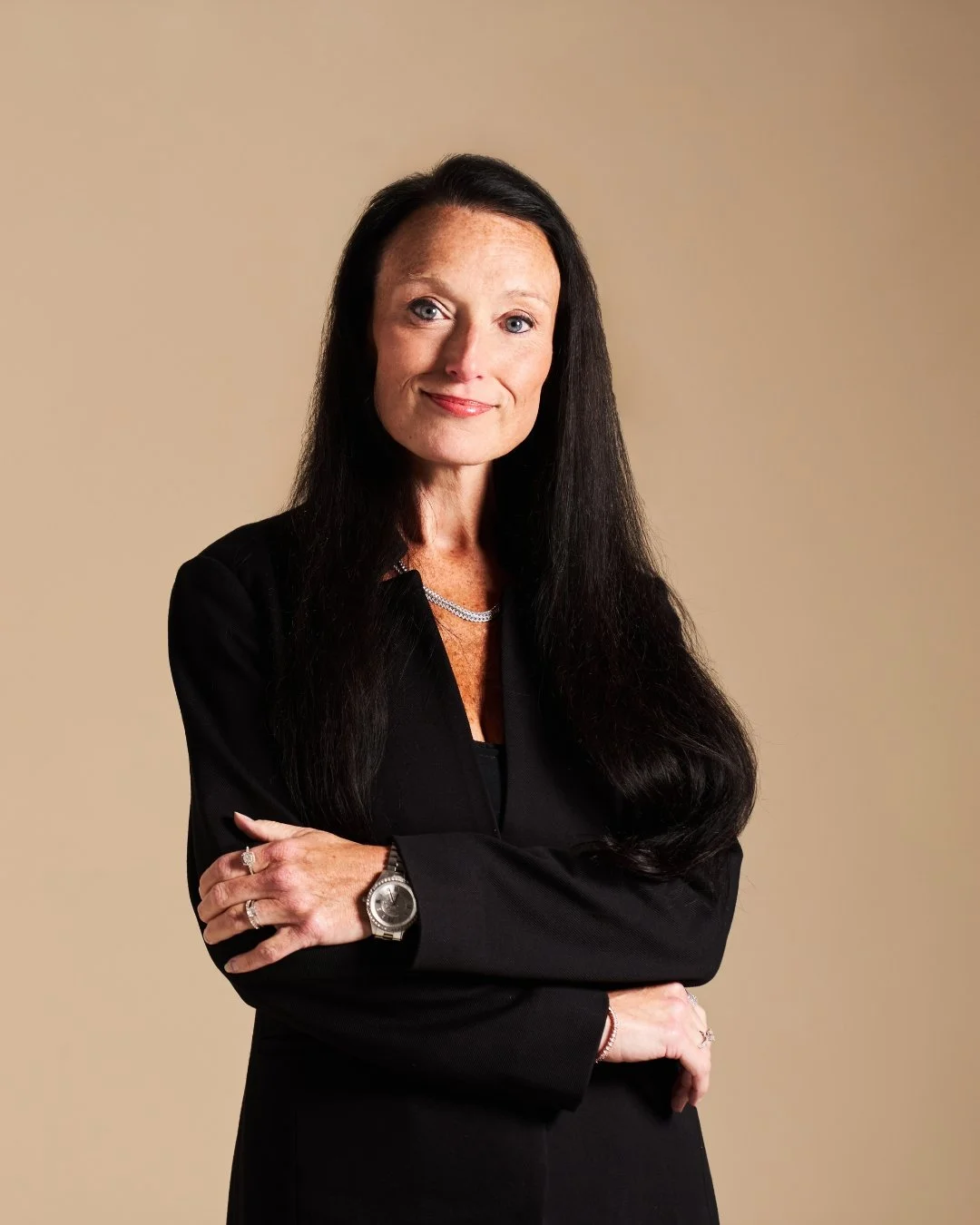 Amy B. RR Living - A woman with long black hair wearing a black blazer, a silver necklace, and a watch, standing with arms crossed against a beige background.