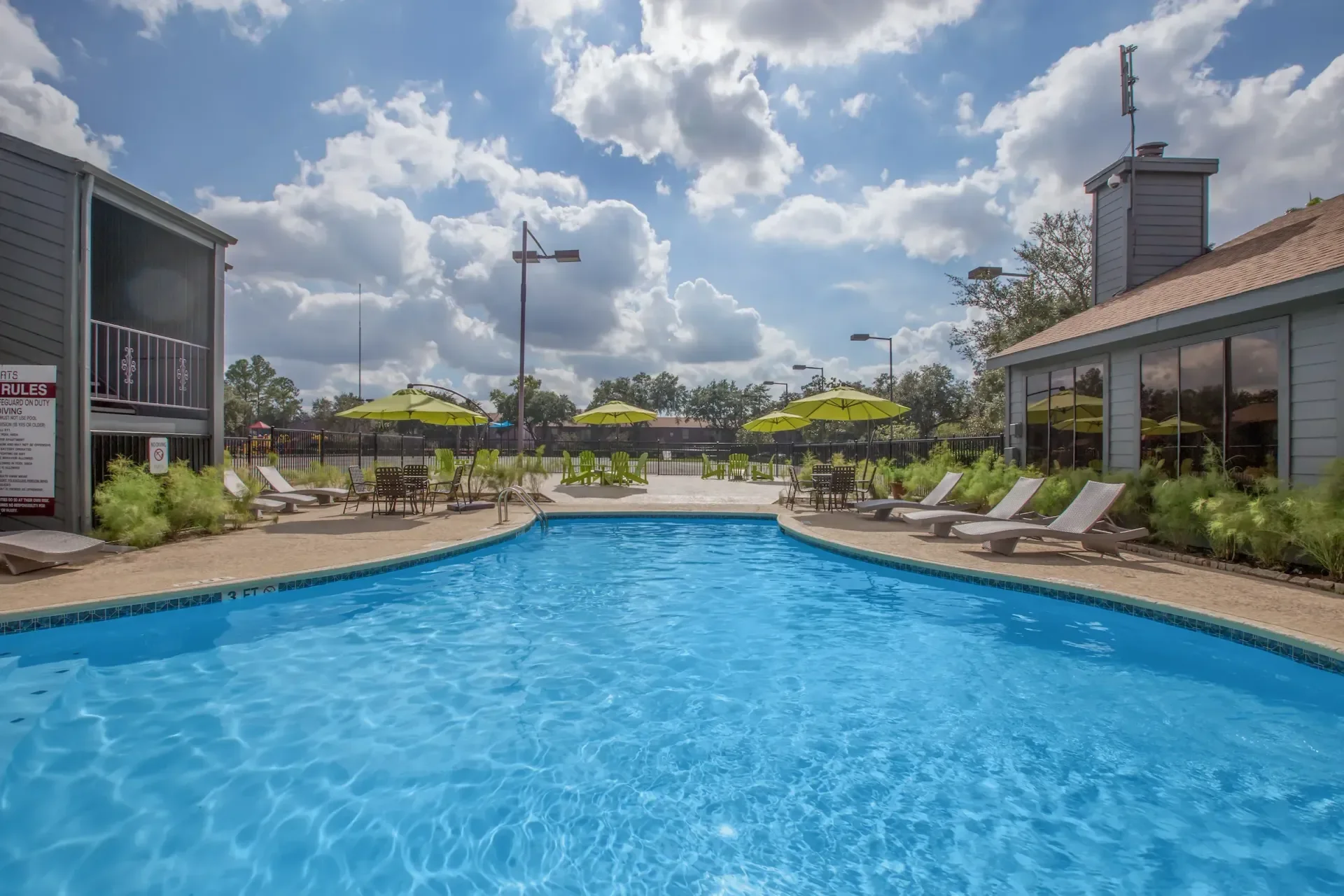 Outdoor swimming pool area with blue water, surrounded by lounge chairs, tables with yellow umbrellas, and a building with large windows. The sky is partly cloudy with the sun shining.