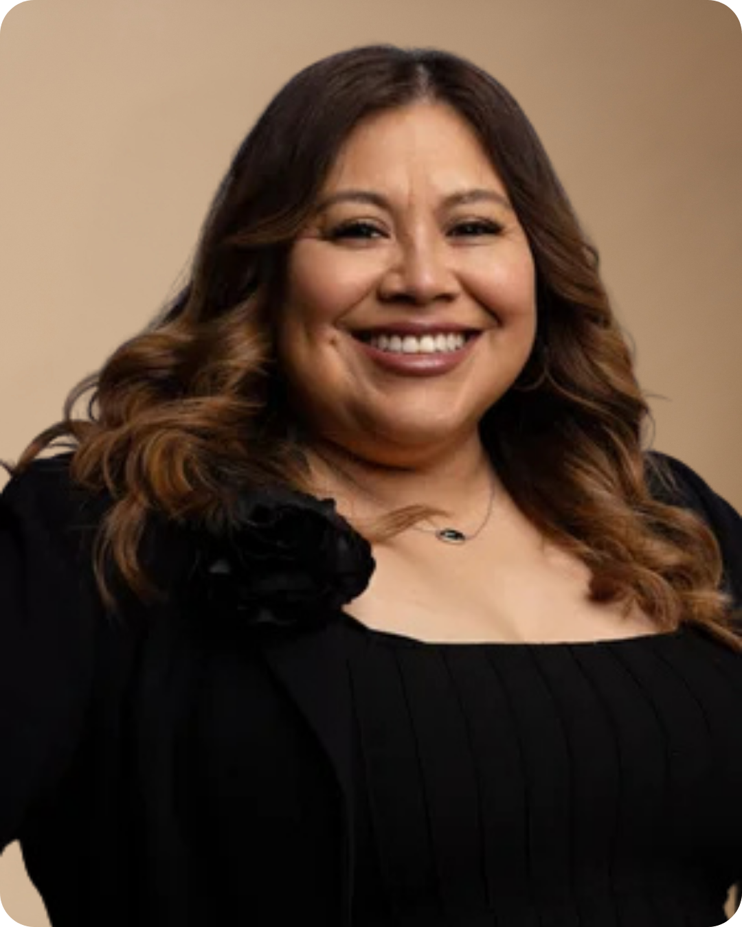 Smiling woman with long wavy brown hair wearing a black top and a black flower accessory on the shoulder.