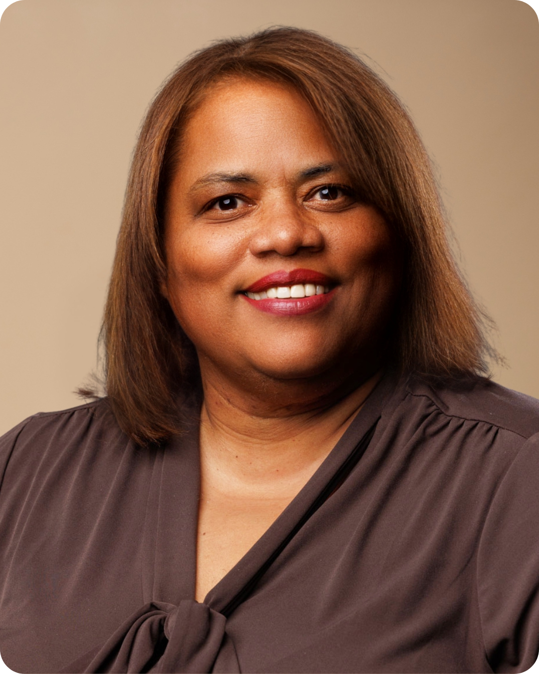 Close-up portrait of a smiling woman with shoulder-length brown hair, wearing a brown blouse with a bow at the neck.