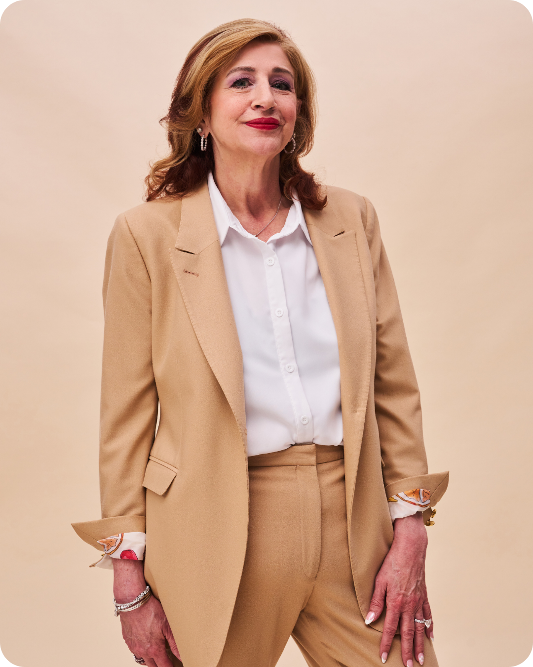 A woman with red hair dressed in a beige suit and white blouse, standing against a plain beige background.