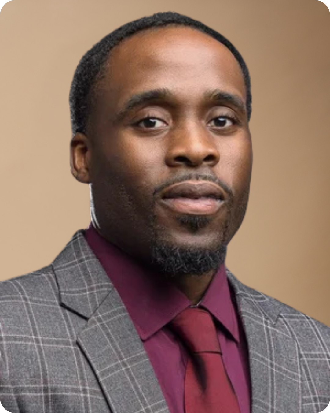 A man with dark hair and a beard wearing a checkered suit, maroon shirt, and maroon tie against a plain background.