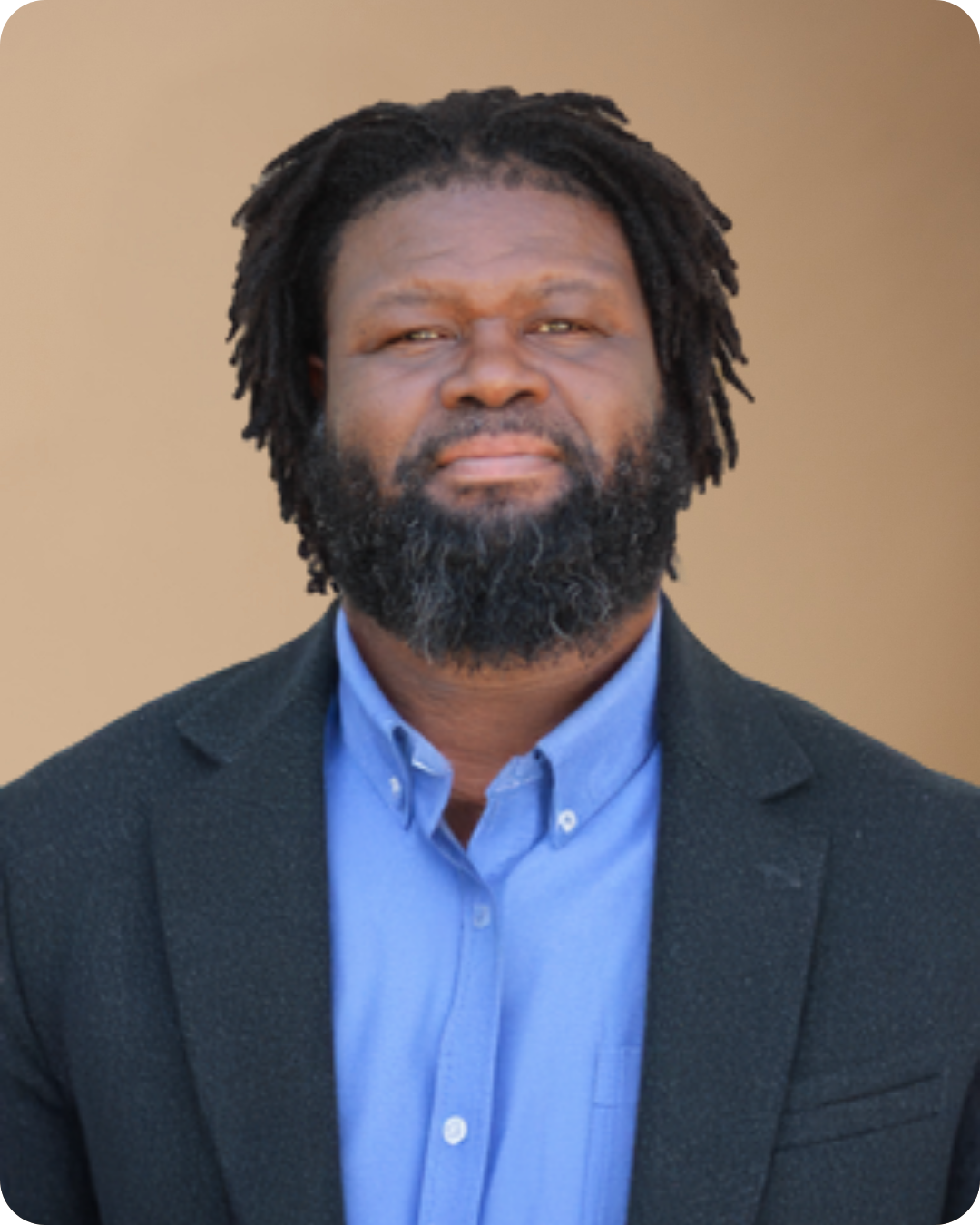 Headshot of a man with dreadlocks and a beard, wearing a blue collared shirt and black blazer, standing outdoors against a beige background.