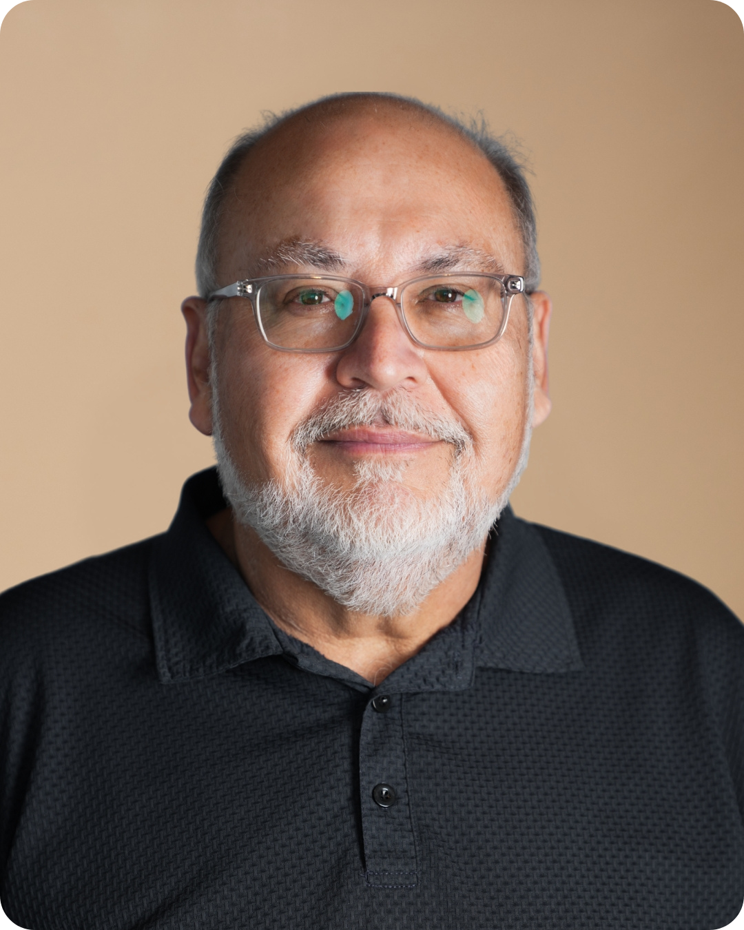Close-up portrait of a senior man with glasses, a gray beard, and a black polo shirt, smiling softly against a plain beige background.