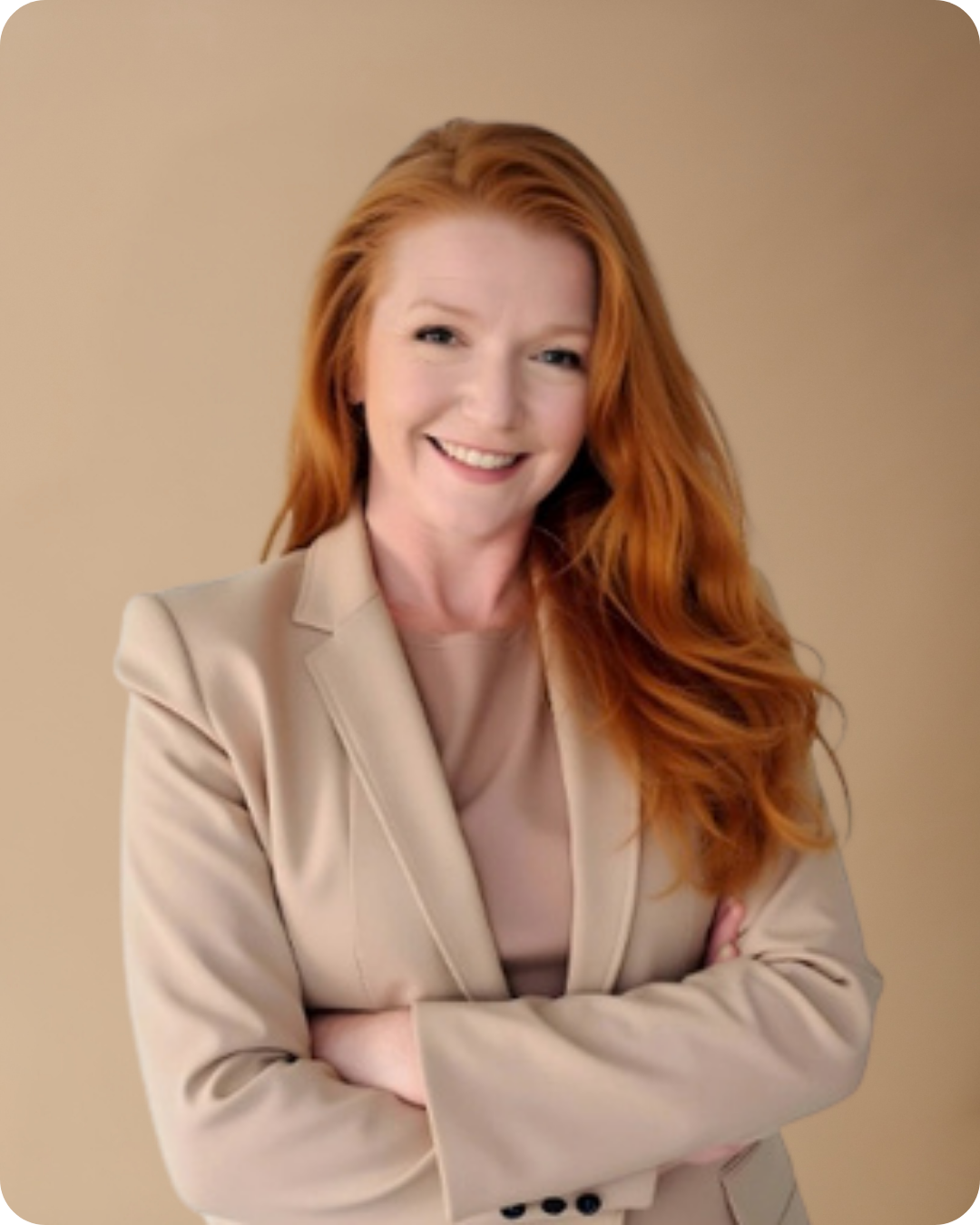 A woman with long red hair smiling and crossing her arms, wearing a beige blazer and top, standing against a plain beige background.