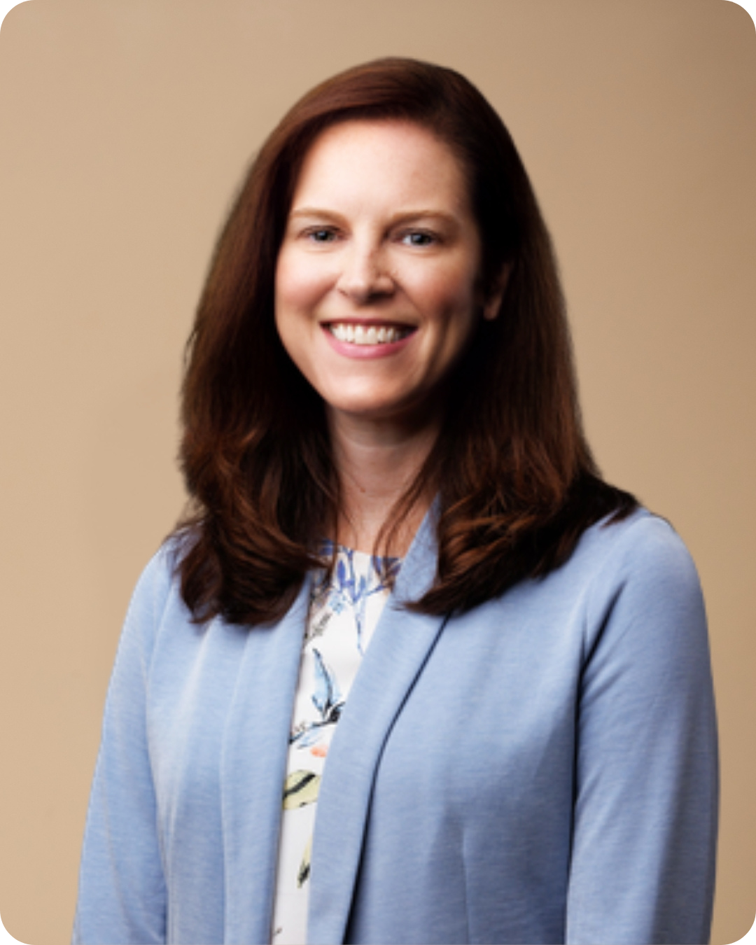 A professional woman with shoulder-length brown hair, wearing a light blue blazer and a floral blouse, smiling against a beige background.