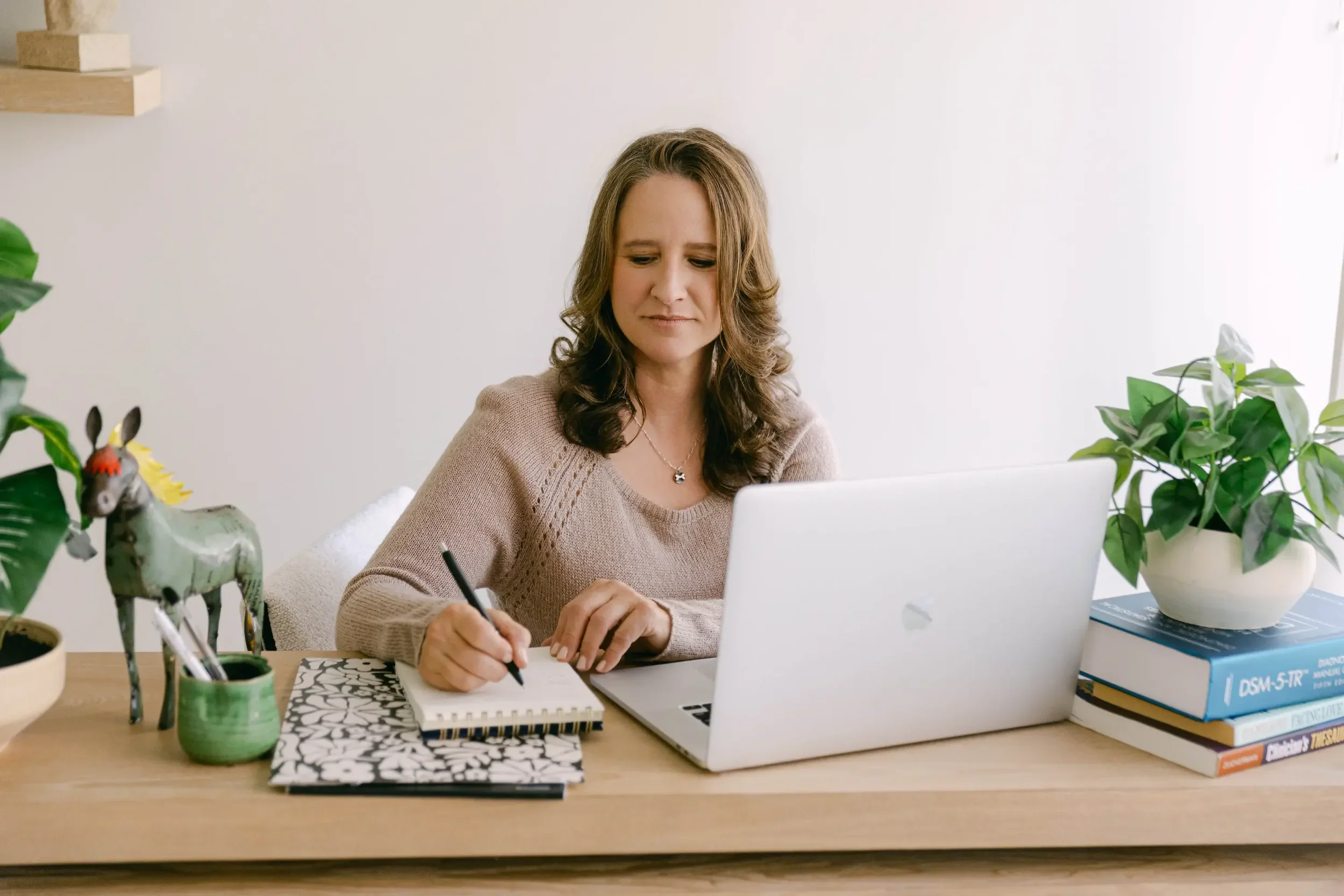 Woman sitting at a desk with a laptop, taking notes in a notebook, surrounded by potted plants and books.