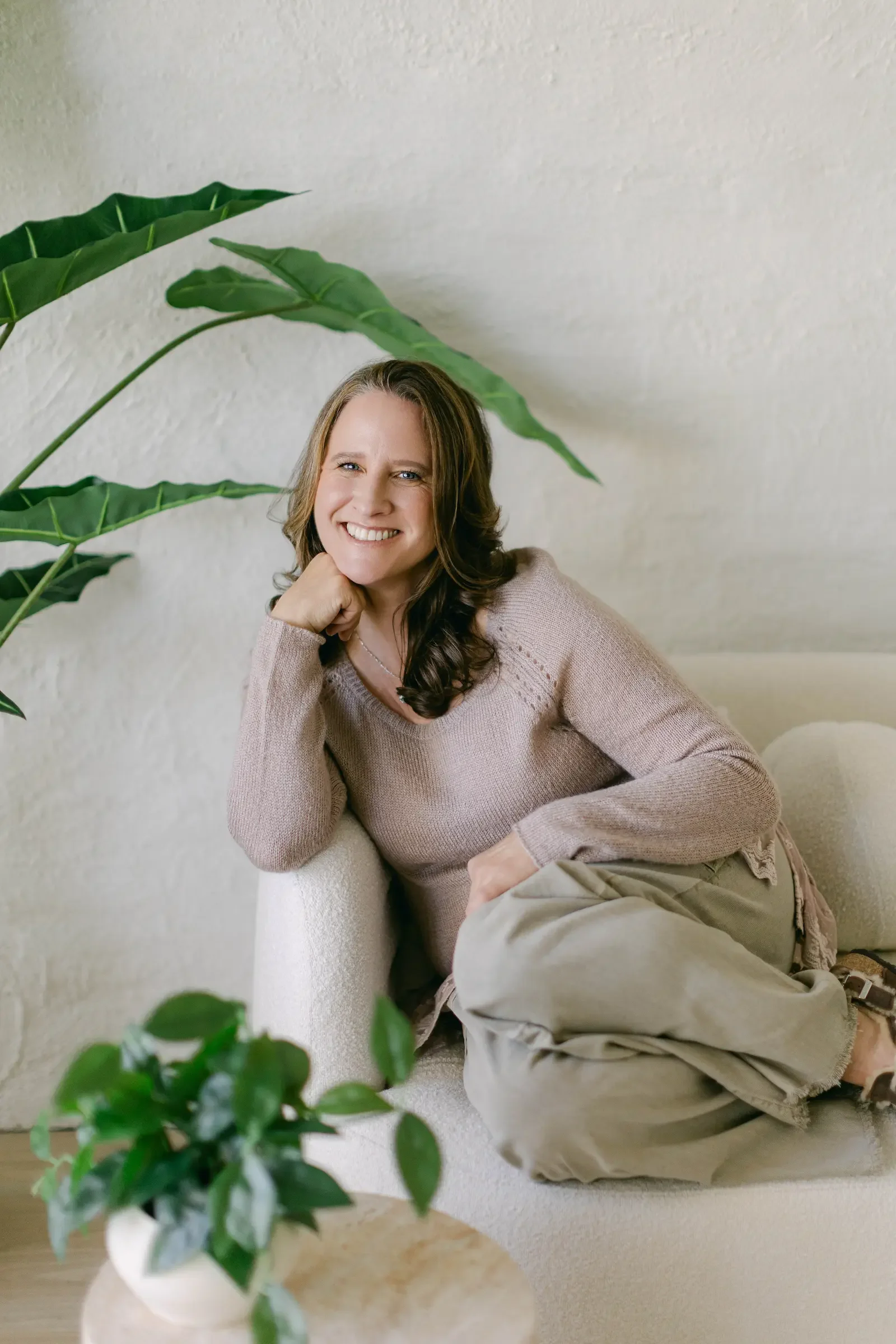 A woman with brown hair, wearing a beige sweater and khaki pants, is sitting on a light-colored sofa, smiling with her head resting on her hand. There are large green plants in the background, and a small green plant in a white pot on a wooden table in the foreground.