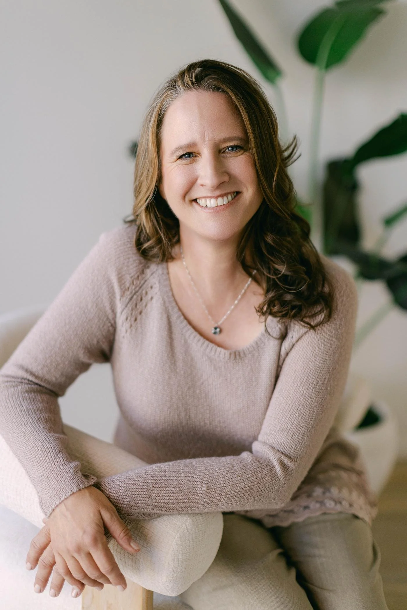 A smiling woman with brown wavy hair, wearing a beige sweater and a necklace, sitting on a chair with a large green plant in the background.