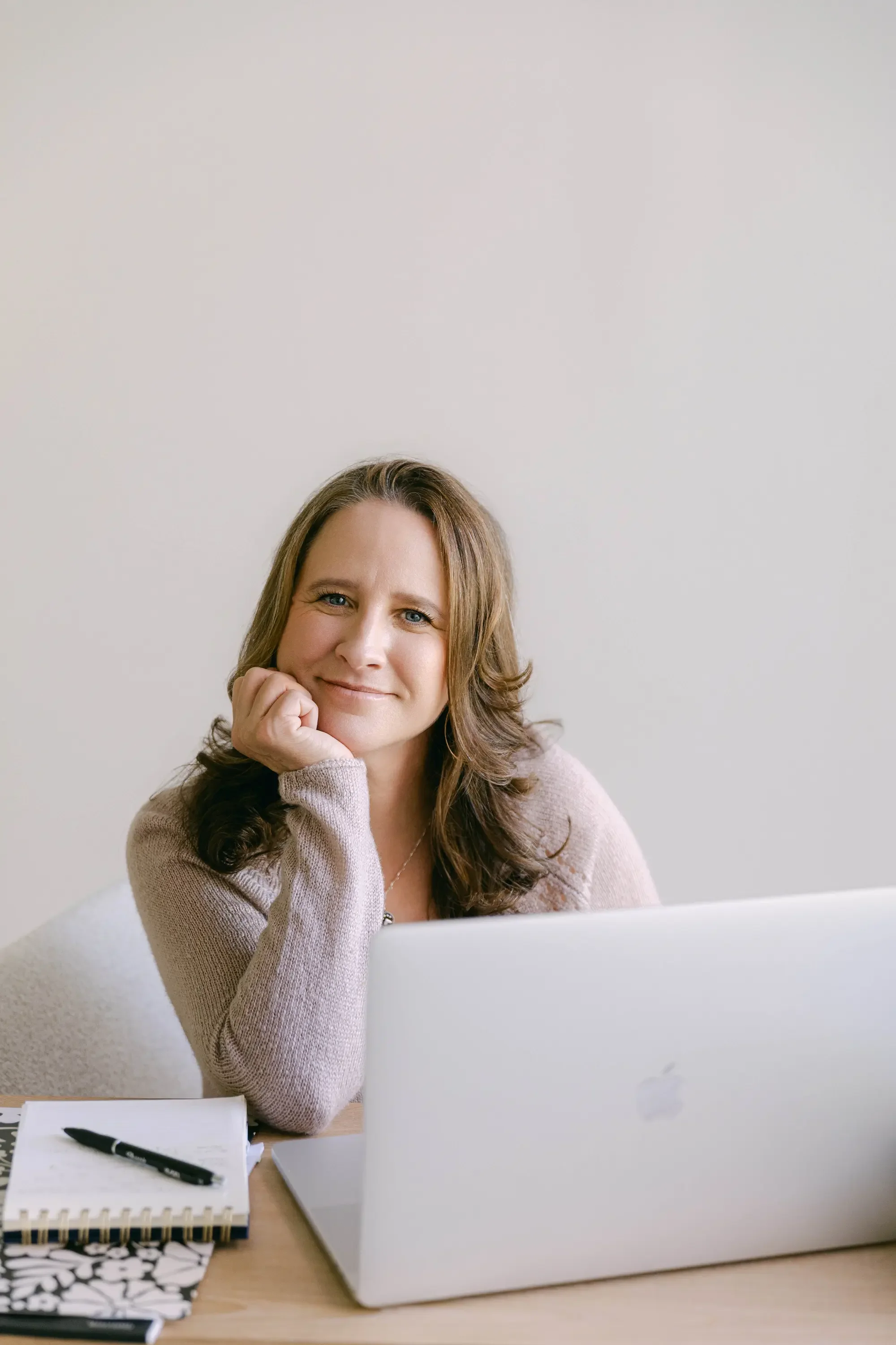 A woman with long wavy brown hair and light skin is sitting at a desk with a silver laptop in front of her. She is smiling, resting her chin on her left hand, and looking at the camera. The desk has a black pen, a notebook, and a black-and-white patterned notepad. The background is a plain white wall.