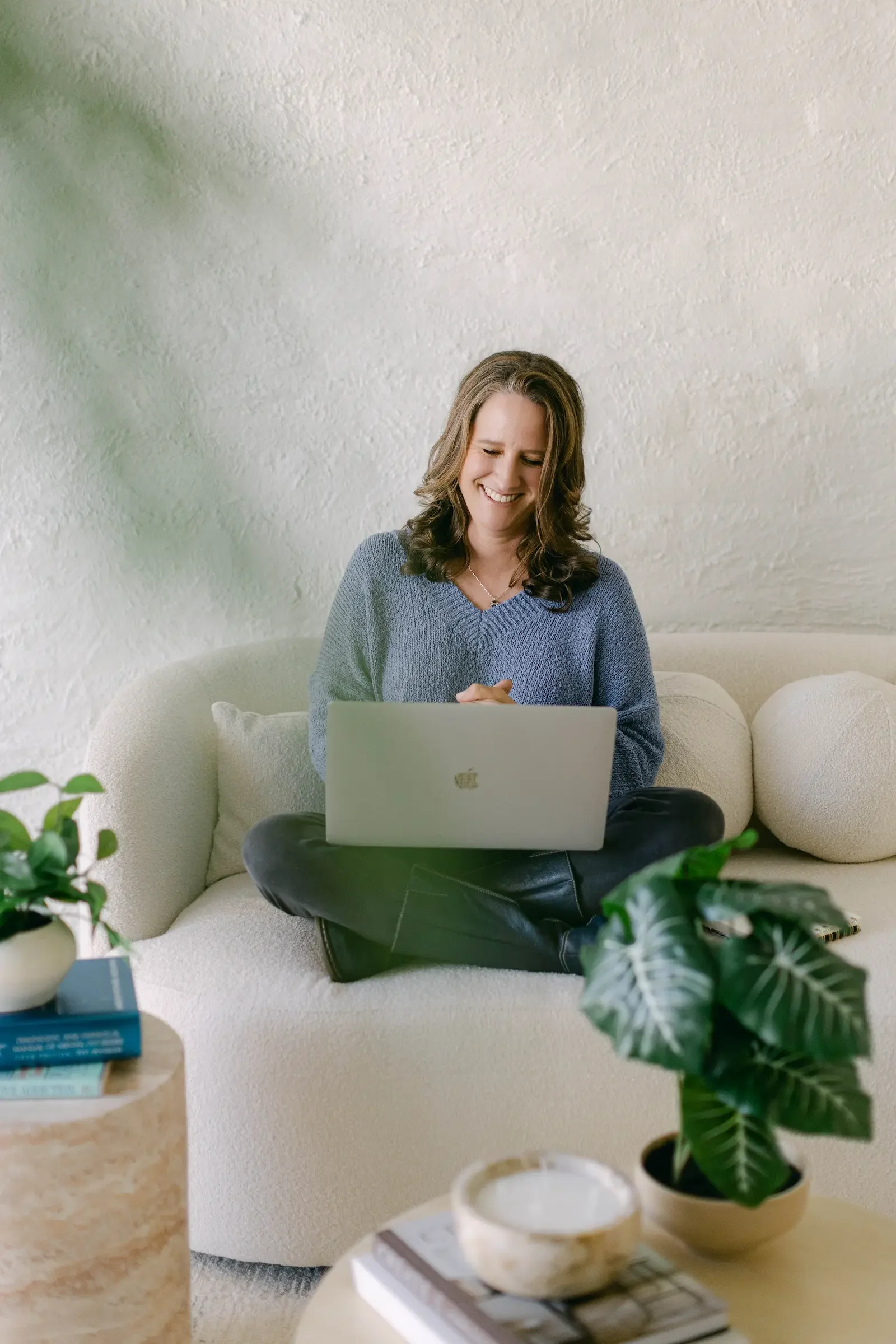 An addiction therapist sitting cross-legged on a white couch, smiling at her laptop, with green plants and books on a wooden table in the foreground.