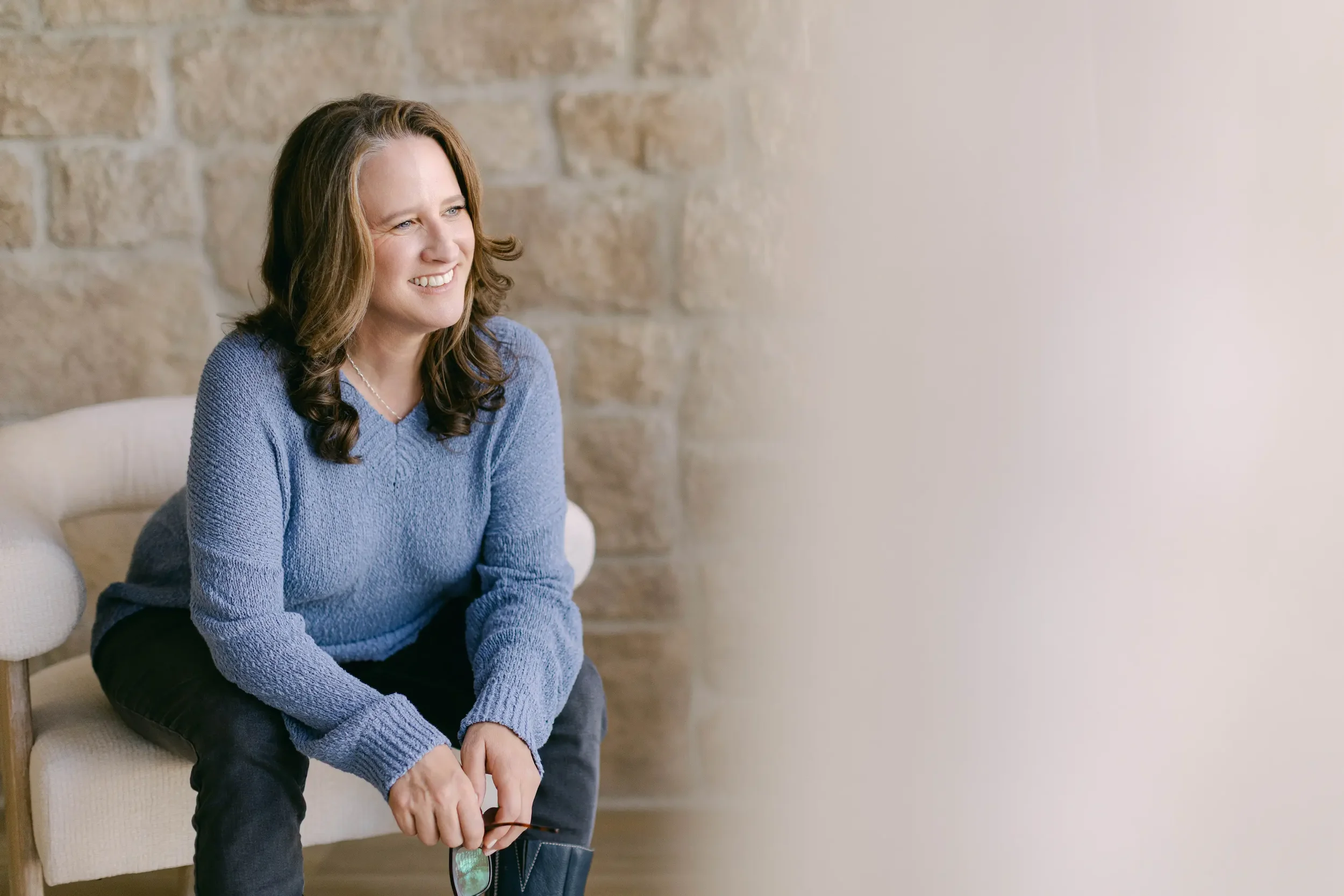 Woman sitting on a sofa, smiling, in a cozy setting with a stone wall.