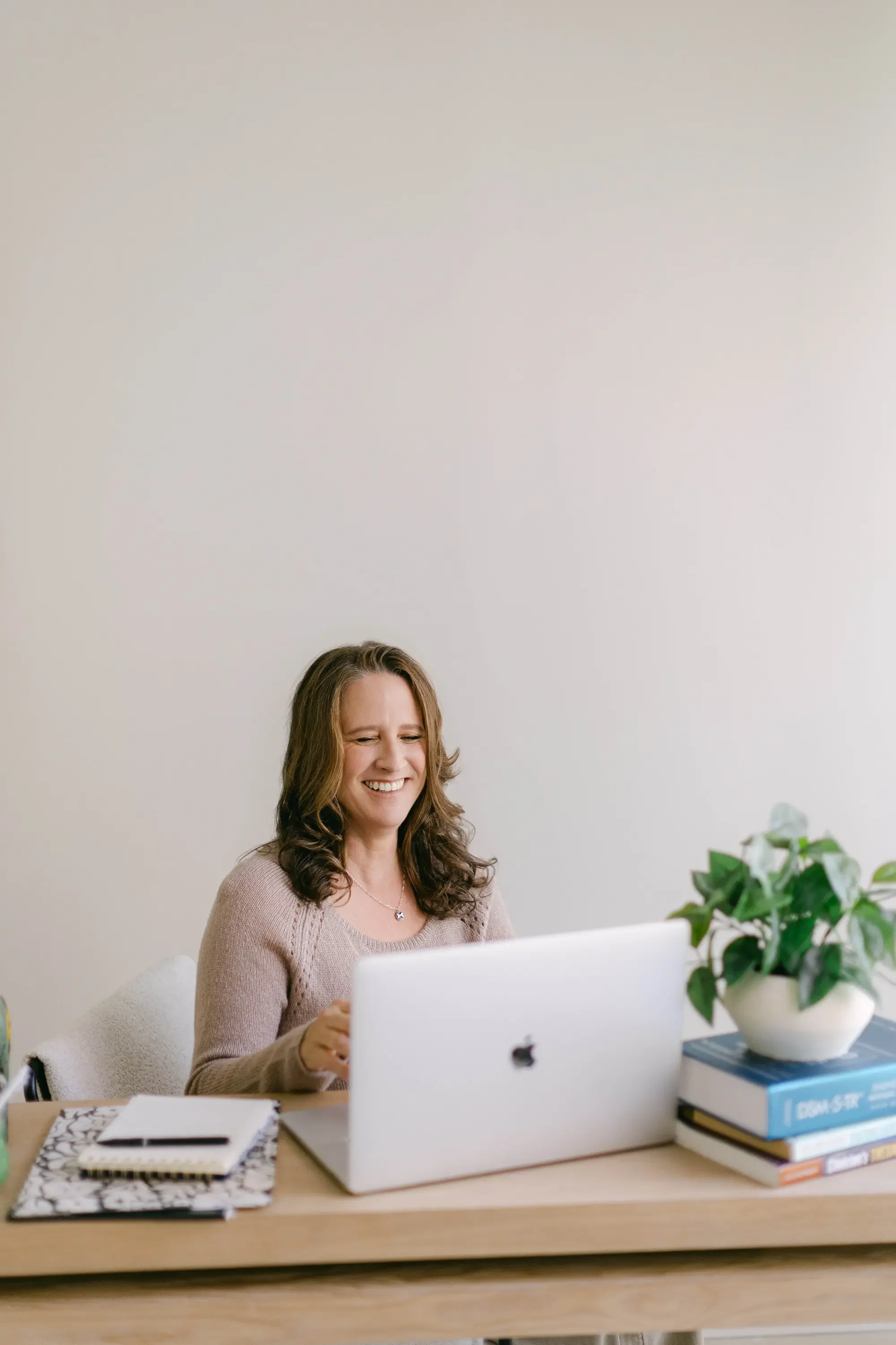 A woman sitting at a desk, smiling while looking at her laptop, with a potted plant and stacked books on her desk.