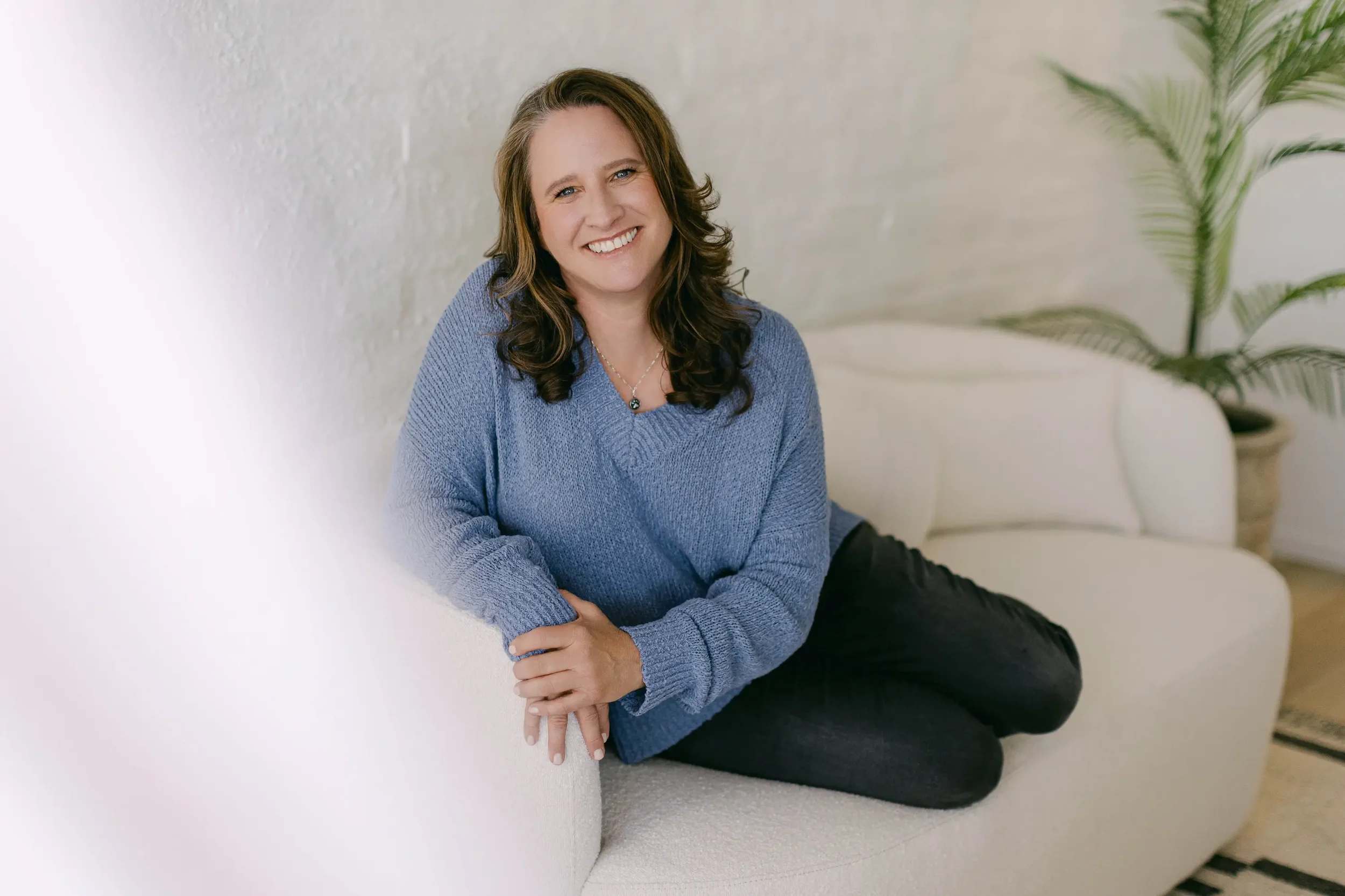 A woman with shoulder-length brown hair wearing a blue sweater and black pants, sitting on a light-colored sofa, smiling, with a large potted plant behind her.