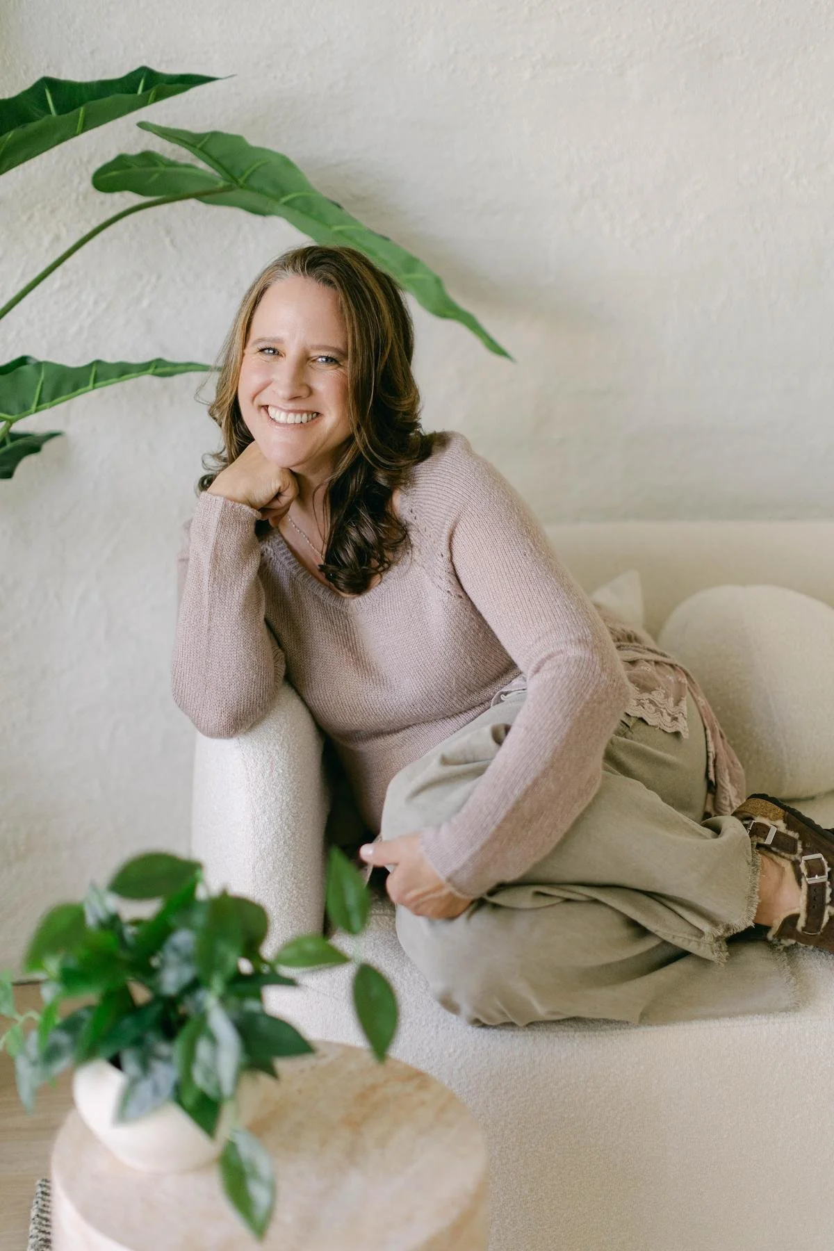 An addiction therapist with long brown hair smiling, sitting on a cream-colored couch with her head resting on her hand, surrounded by green houseplants.