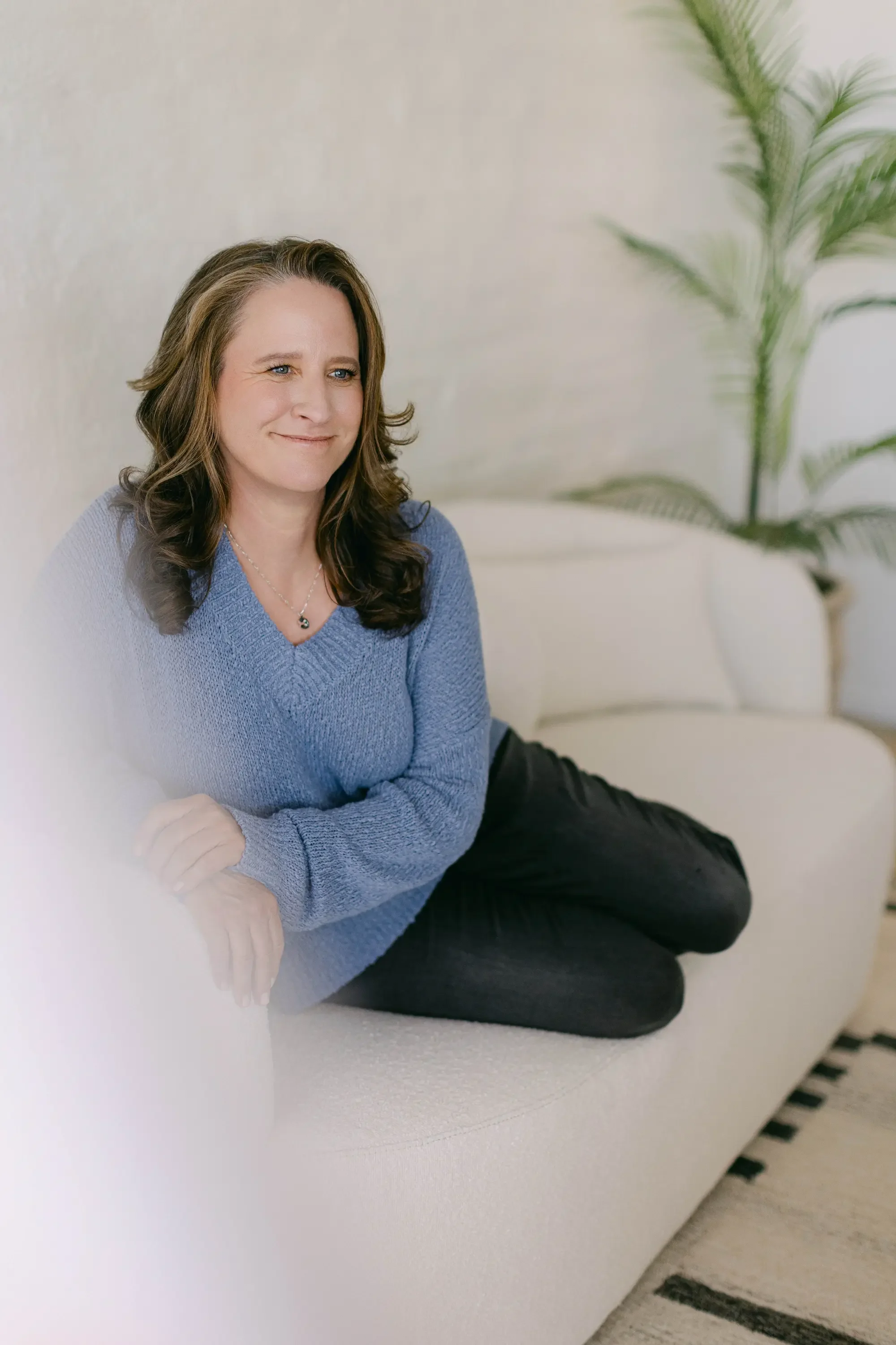 A woman with brown hair sitting on a white sofa, wearing a blue sweater and black pants, smiling, with a green potted plant in the background.