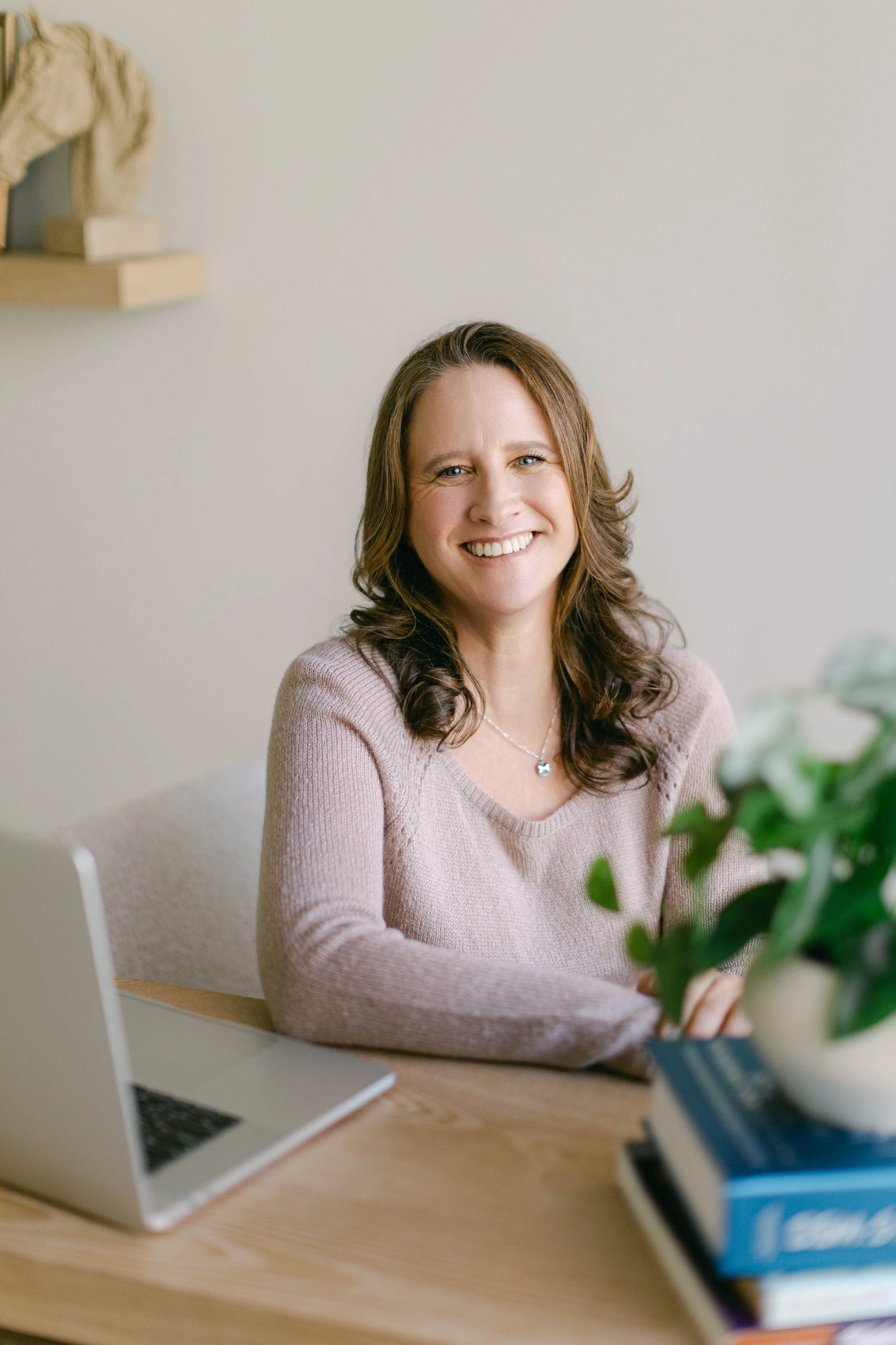 An EMDR therapist with brown hair smiling at the camera, sitting at a desk with a laptop, bookshelf, and potted plant nearby.