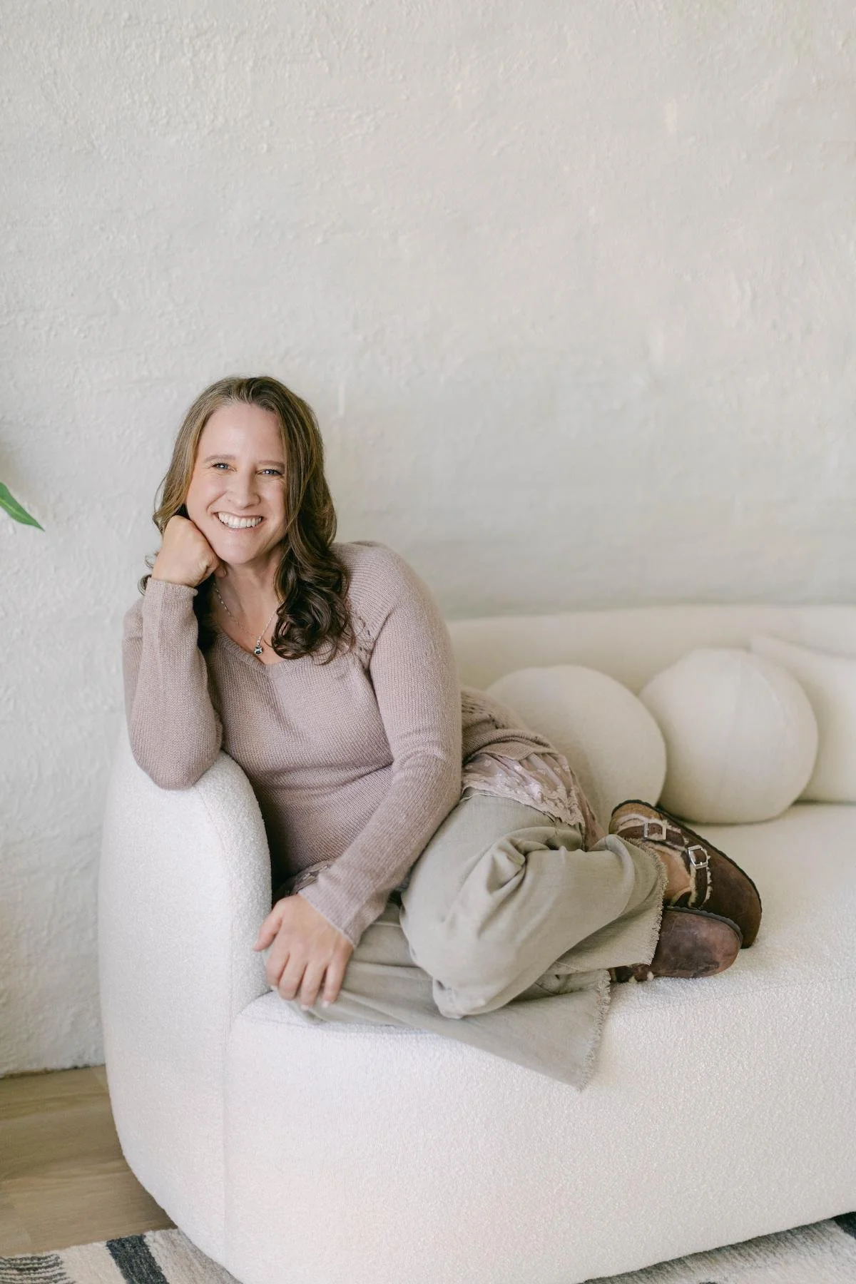 A therapist with long brown hair, smiling, sitting on a white sofa in a cozy room with a textured white wall.