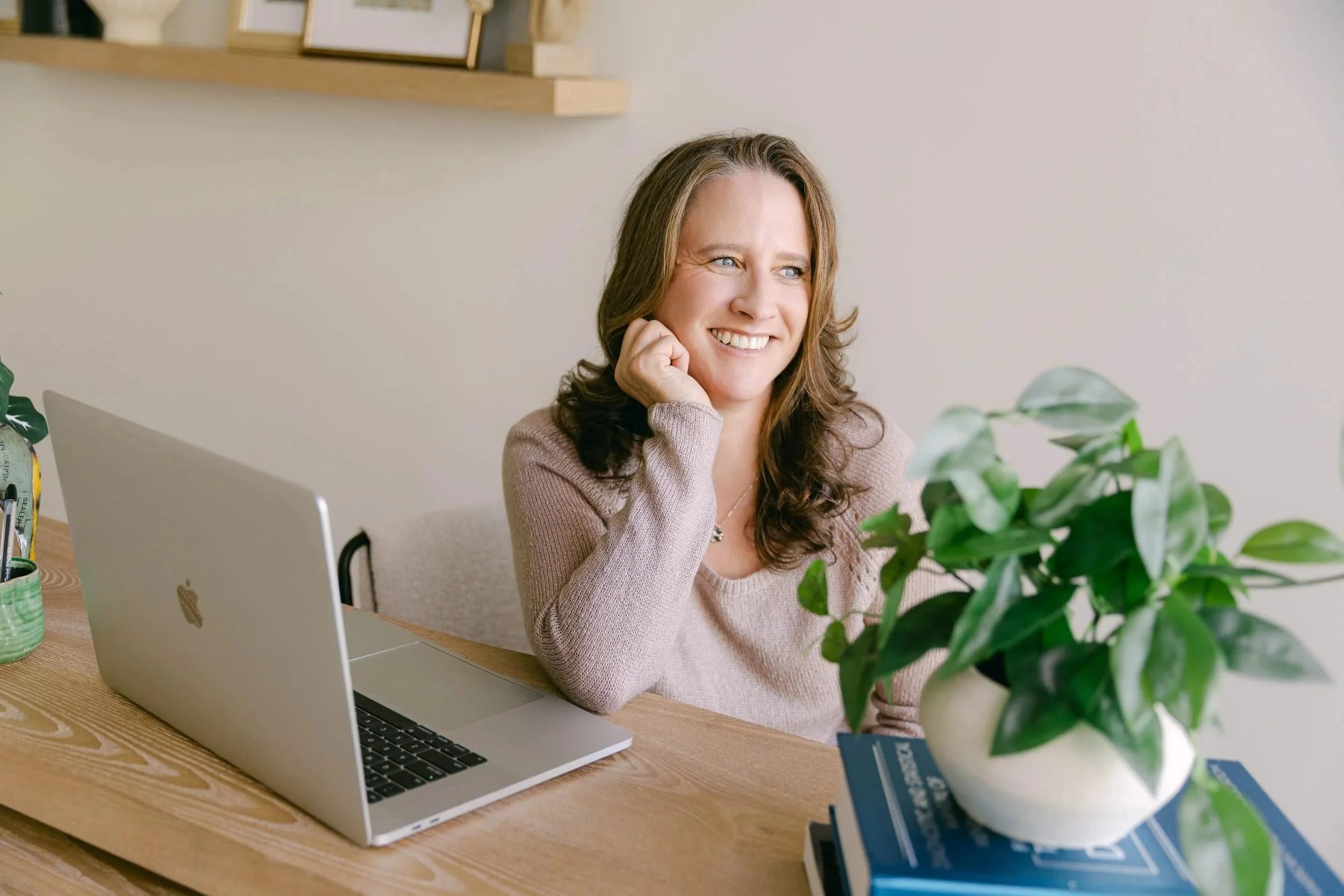 A therapist sitting at a desk with a laptop, smiling and looking away, with a potted plant, books, and a wall shelf in the background.