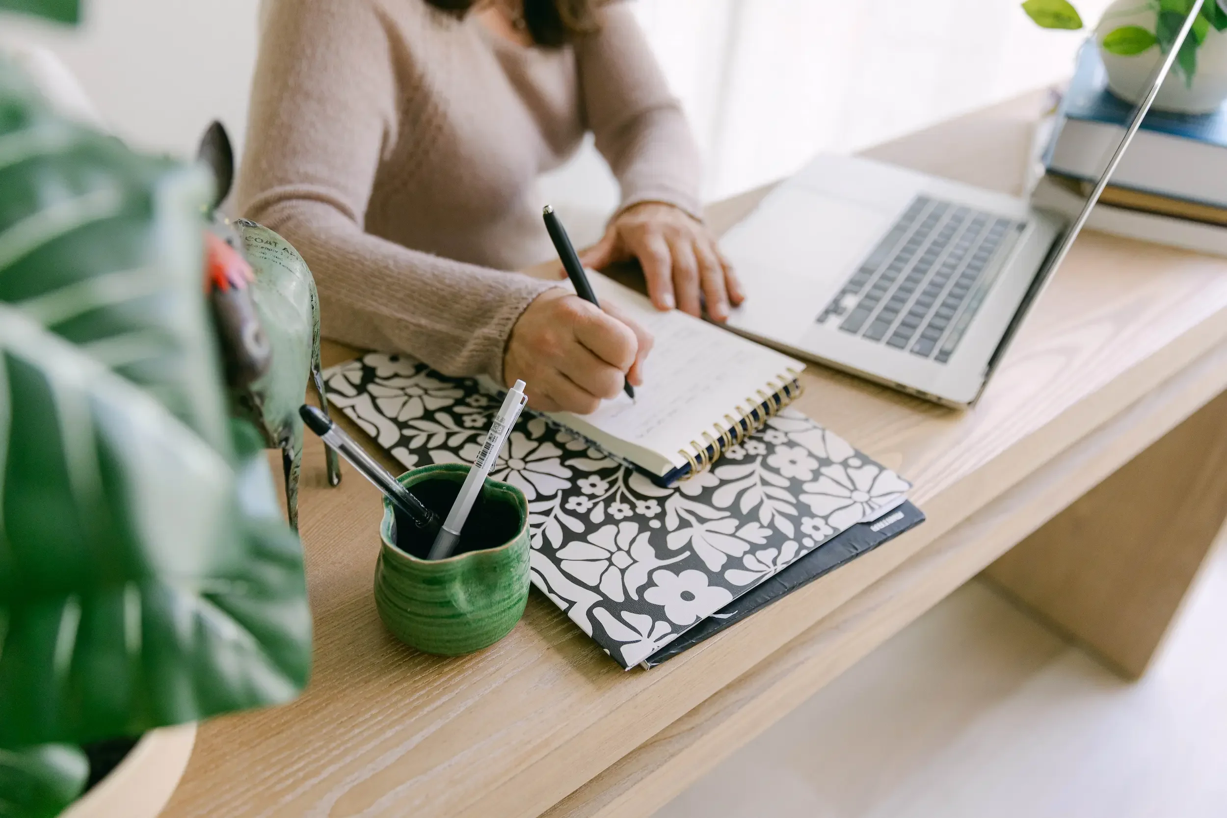 A person sitting at a desk, writing in a notebook with a pen, with a laptop open in front of them. A green potted plant sits to the side, and there are stacked books and a small container with pens or pencils nearby.