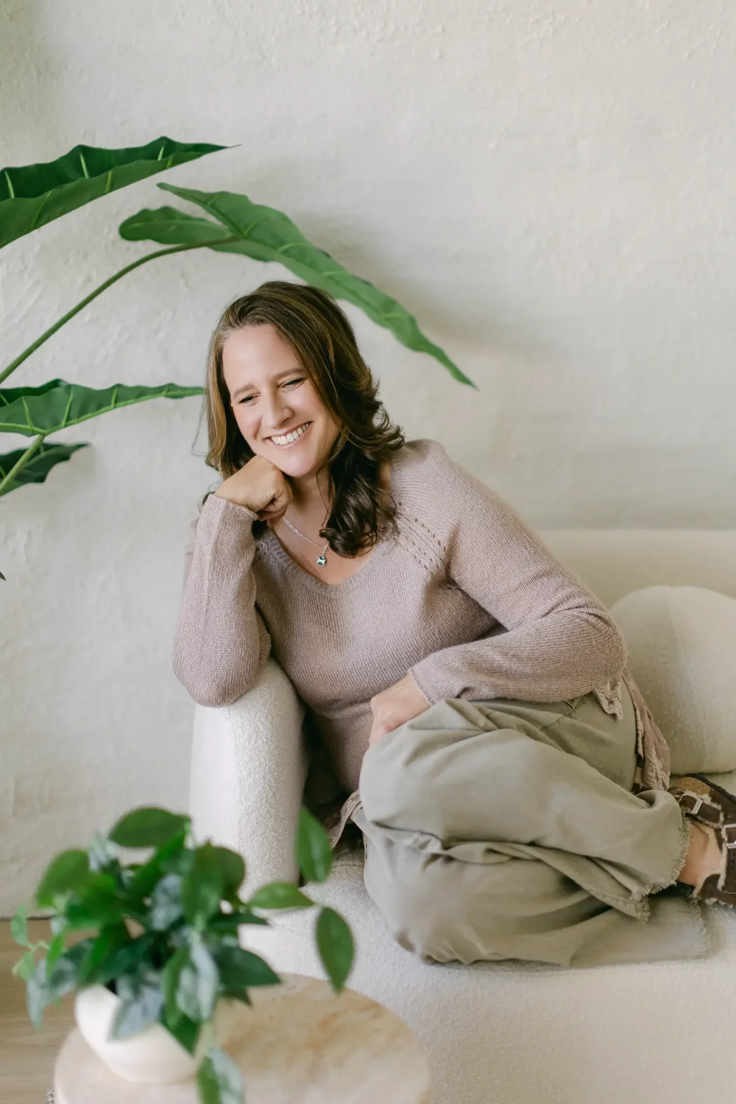 A woman with brown hair sitting on a light-colored sofa, smiling with her eyes closed, leaning her head on her hand, wearing a beige sweater and khaki pants, in a cozy room with green plants and a white textured wall.