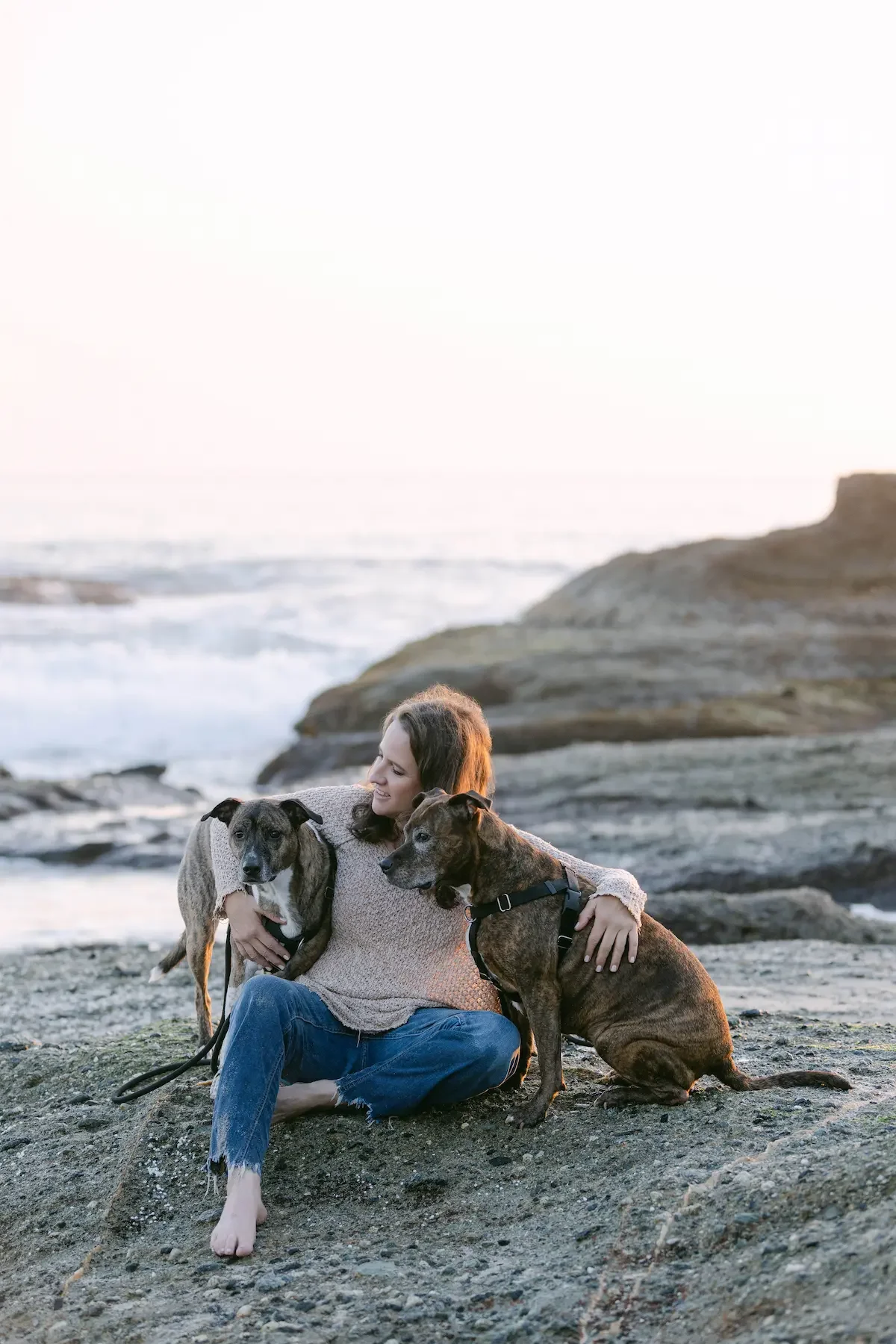 A therapist sitting on a rocky beach with two dogs, one on each side, during sunset.