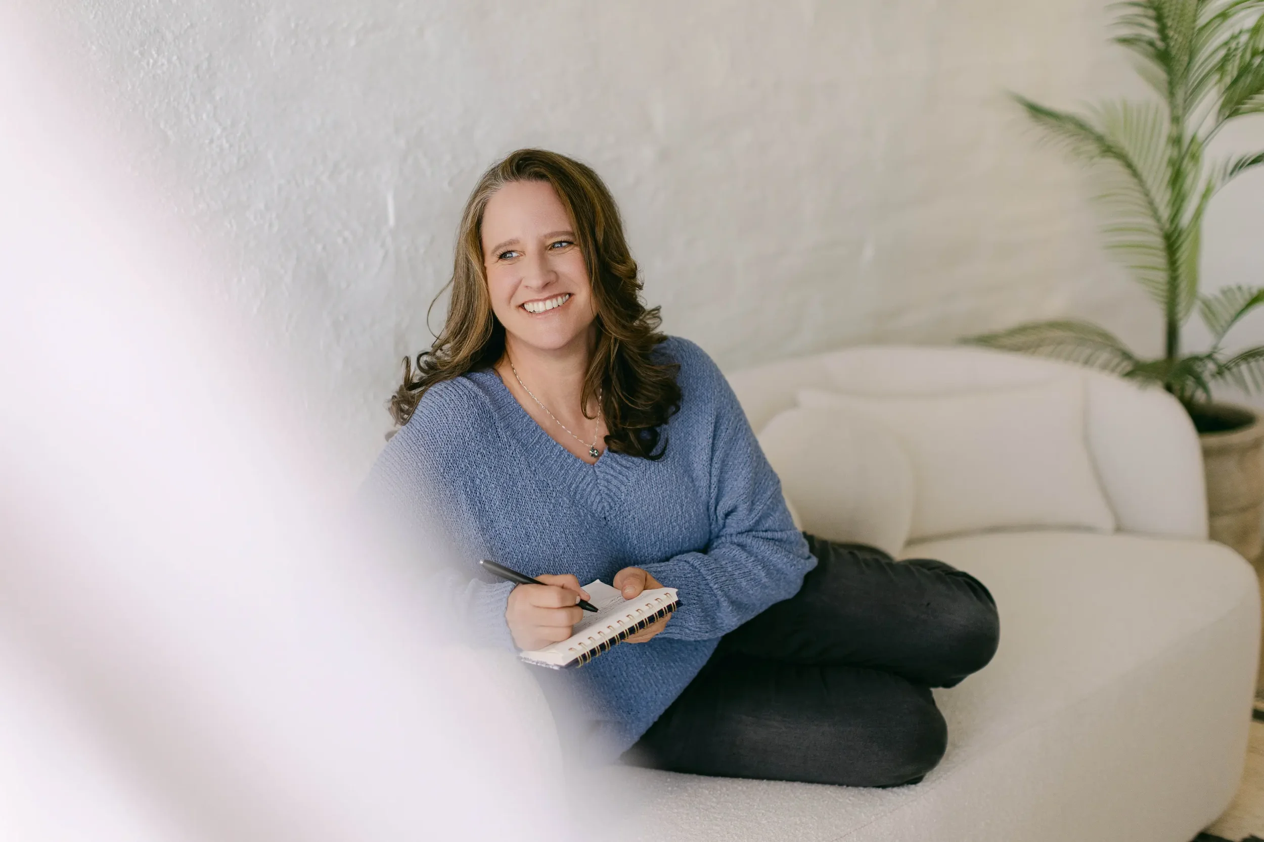 A woman with brown hair wearing a blue sweater, sitting casually on a white couch, smiling, holding a notepad and pen, with a potted plant in the background.