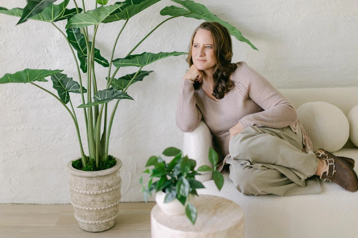 Addiction counselor with dark brown hair, blue eyes, and wearing a tan sweater sitting on an ivory couch with her head resting on her right hand as she looks off to the side. A large green plant is off to the left and a smaller green plant in front.