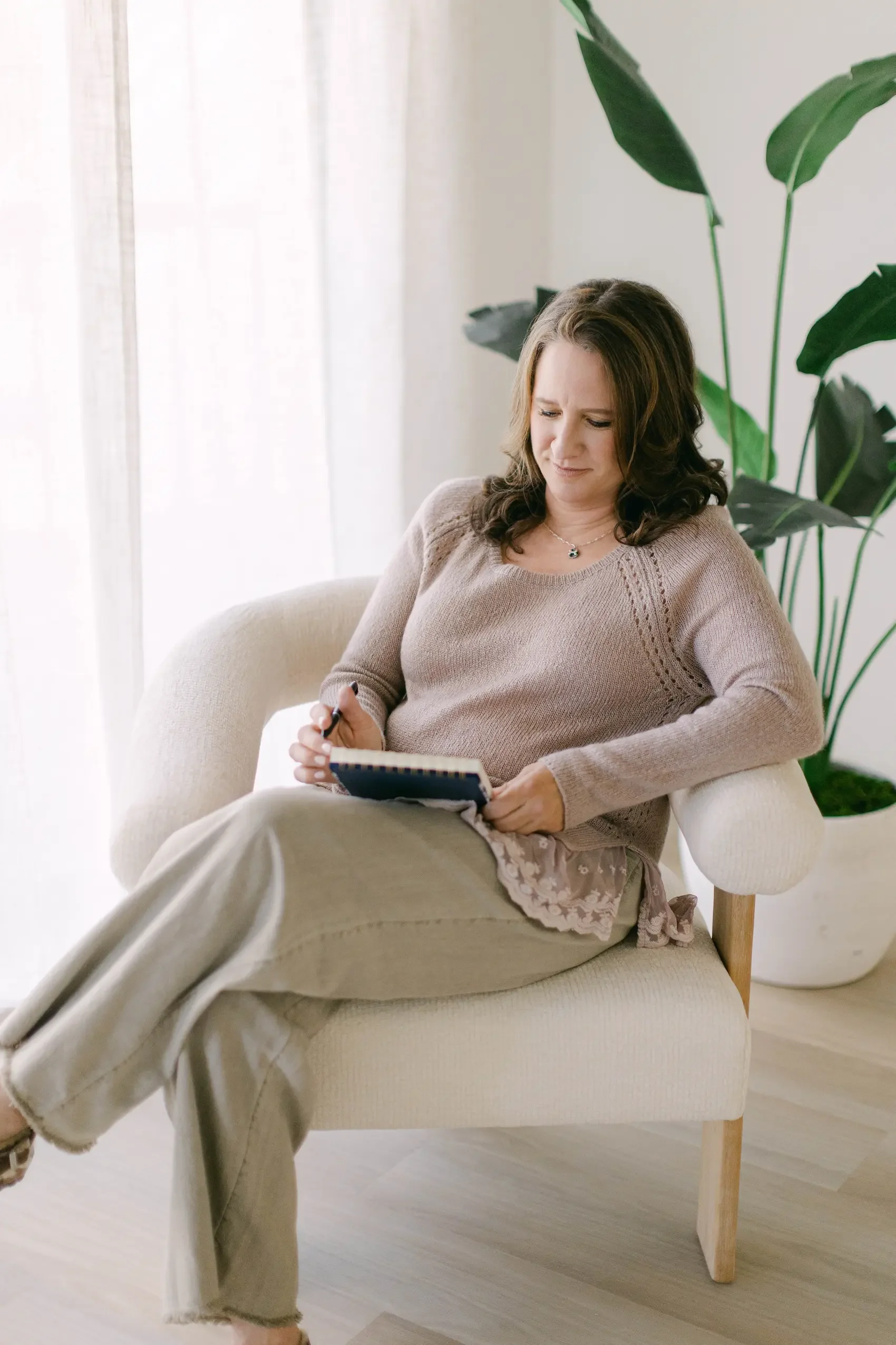 A woman sitting on a light-colored armchair, holding a notebook and pen, with large green plants and a light-colored curtain behind her in a bright, modern room.