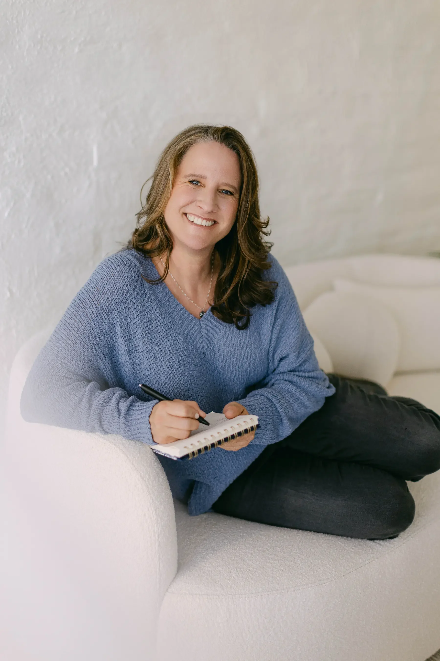 An EMDR and addiction therapist sitting on a white couch, smiling, holding a pen and a notebook, wearing a blue sweater and black pants.