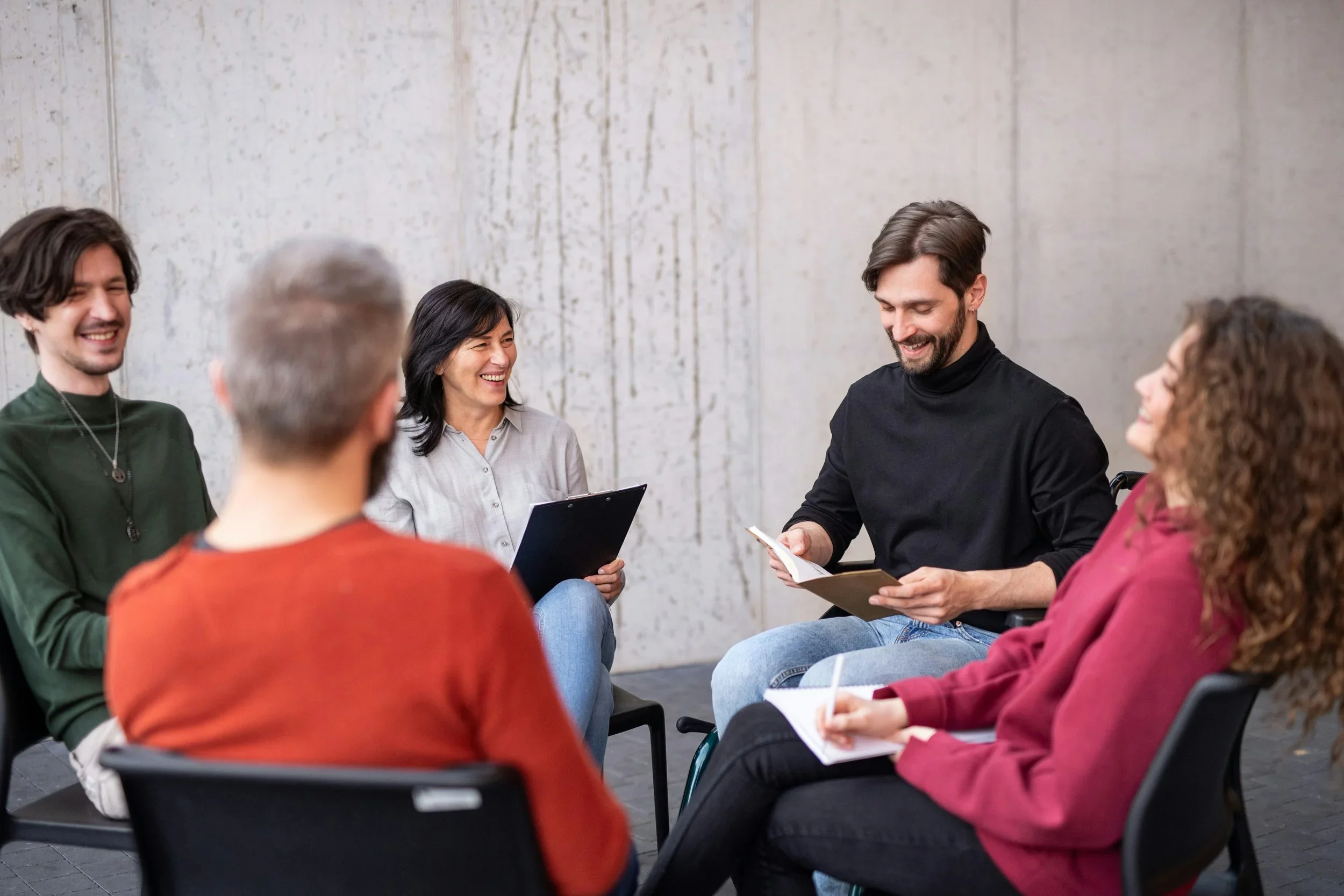 A group of five people sitting in a circle, smiling and having a discussion in a modern room with concrete walls.