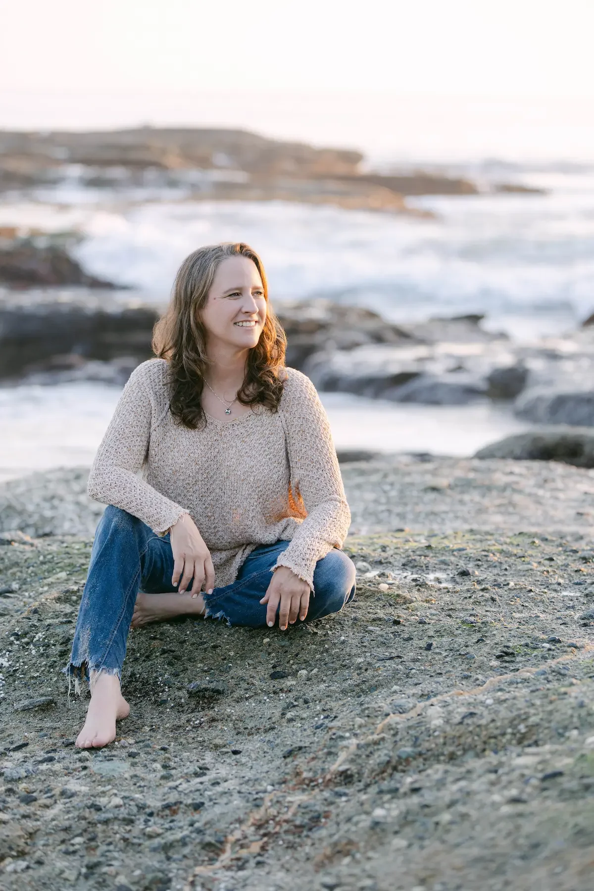 An addiction therapist sitting on a rocky beach with ocean waves in the background, smiling and looking to the side.