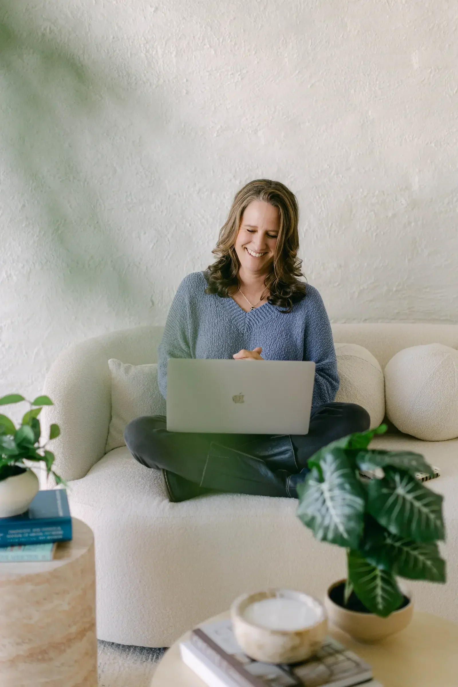A woman sitting on a beige sofa working on a laptop, surrounded by houseplants on the coffee table in the foreground.