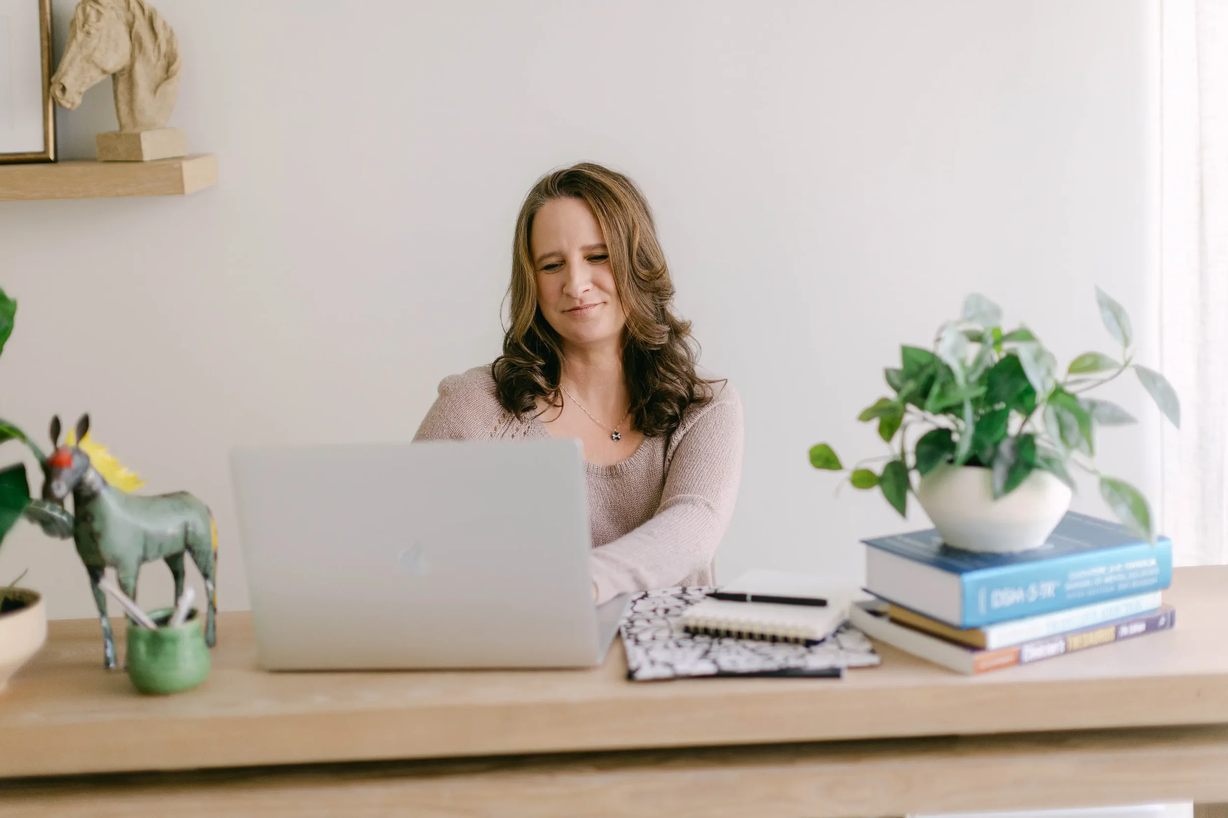 A woman sitting at a desk with a laptop, notebooks, and a pen. There are green plants and decorative items on the desk and a shelf on the wall behind her.