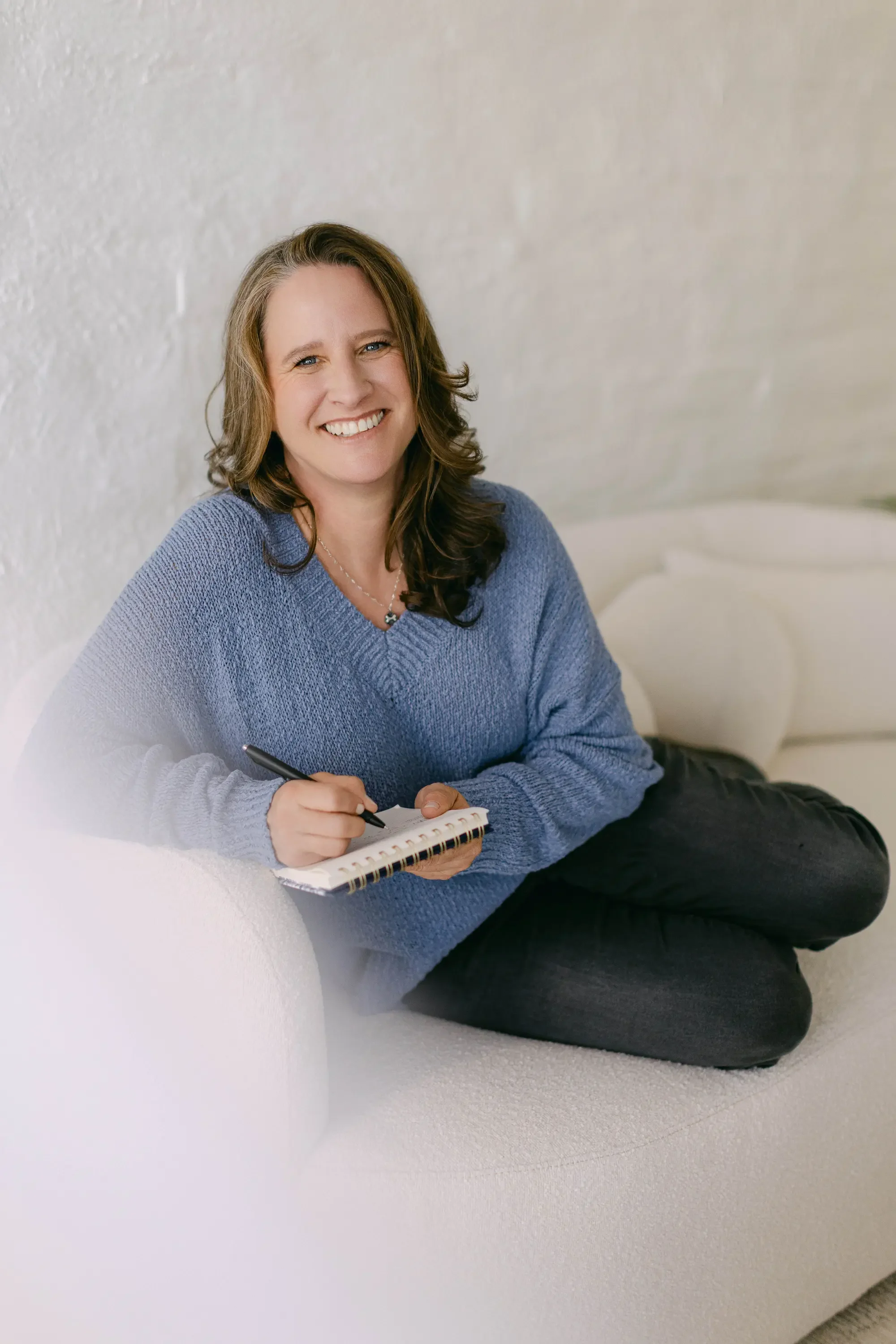 Woman sitting on a white couch, smiling, wearing a blue sweater and black pants, holding a pen and notebook.