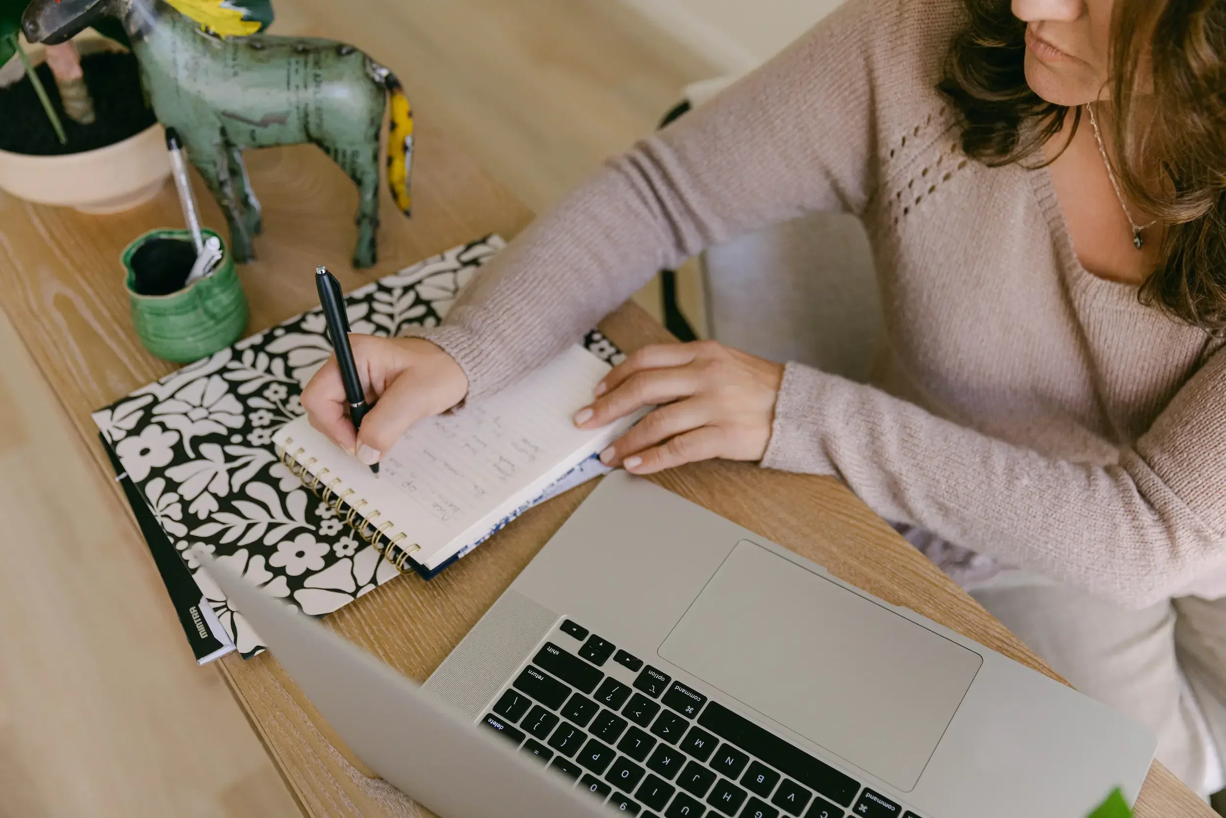 A woman wearing a beige sweater is writing in a notebook at a wooden table. On the table, there is a decorative black and white floral notebook, a mug with a green plant, a small green container, a green painted ceramic horse figurine, and a laptop.