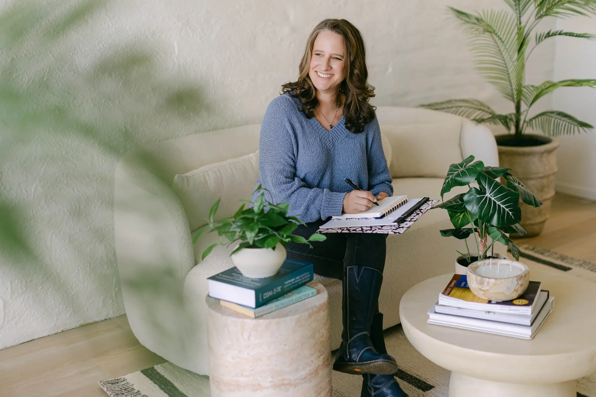 A woman sitting on a beige sofa, smiling with a notebook and pen, surrounded by potted plants and books in a cozy living room.