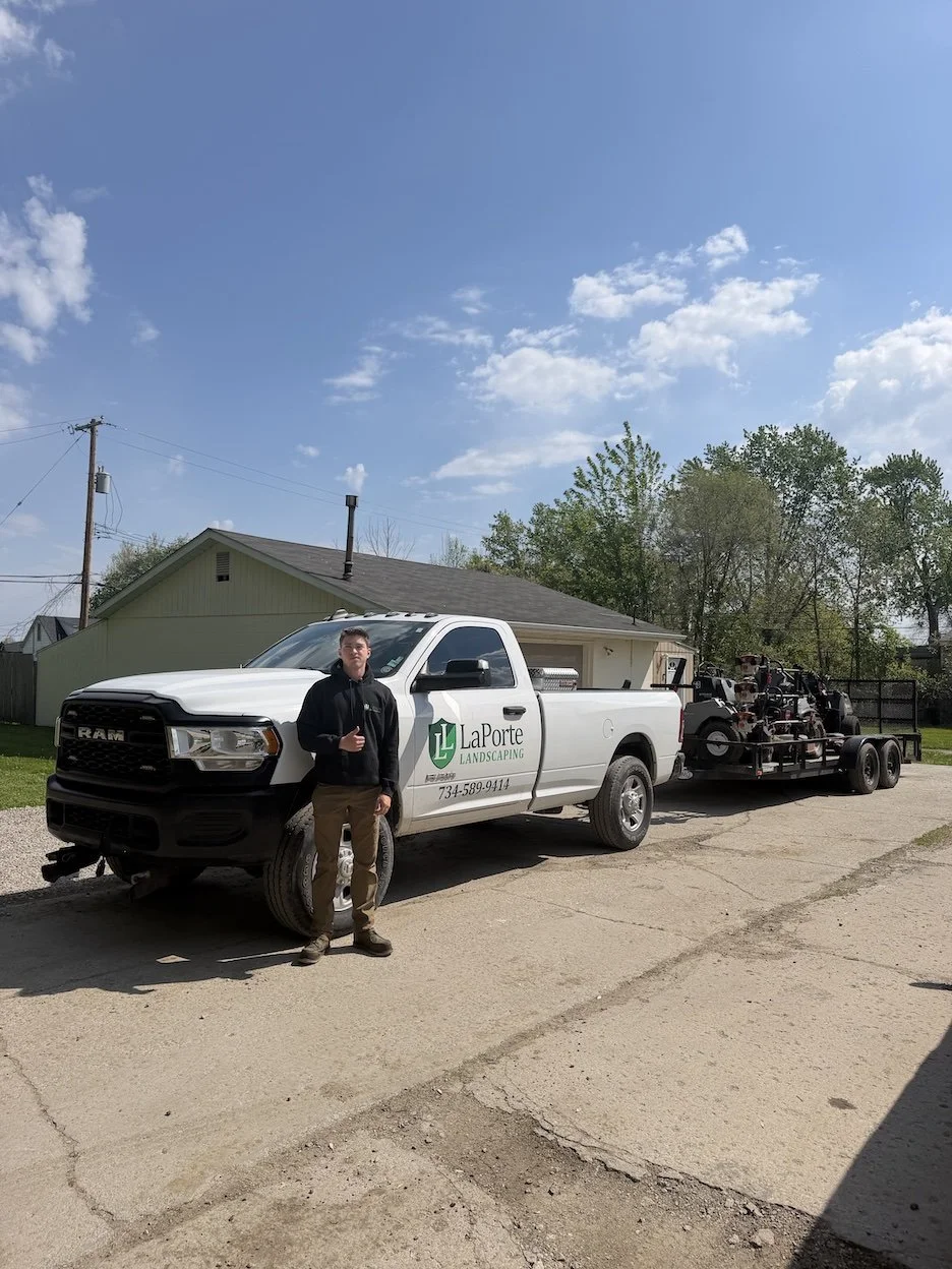 A man standing next to a white pickup truck with a landscaping business logo on the door. The truck is on a driveway with a trailer attached, carrying equipment. The background includes a house, trees, and a partly cloudy sky.