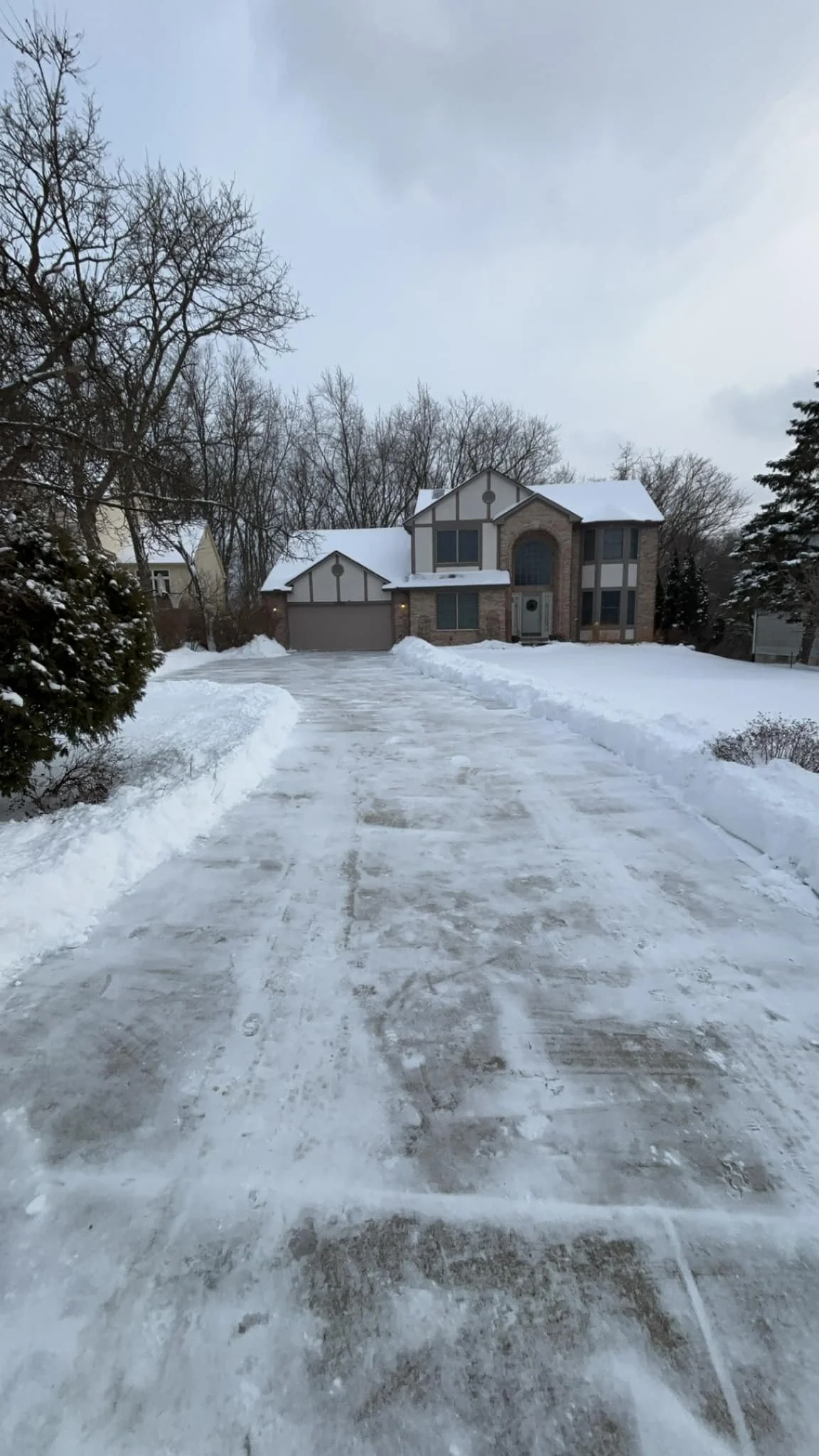 Snow-covered driveway leading to a house with a stone facade, surrounded by leafless trees under an overcast sky.