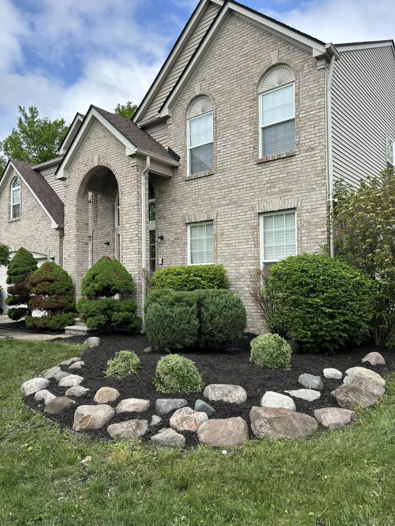 A two-story brick and siding house with arched windows and a front garden. The garden has neatly trimmed bushes, a stone border, and a well-maintained lawn, with a partly cloudy sky in the background.
