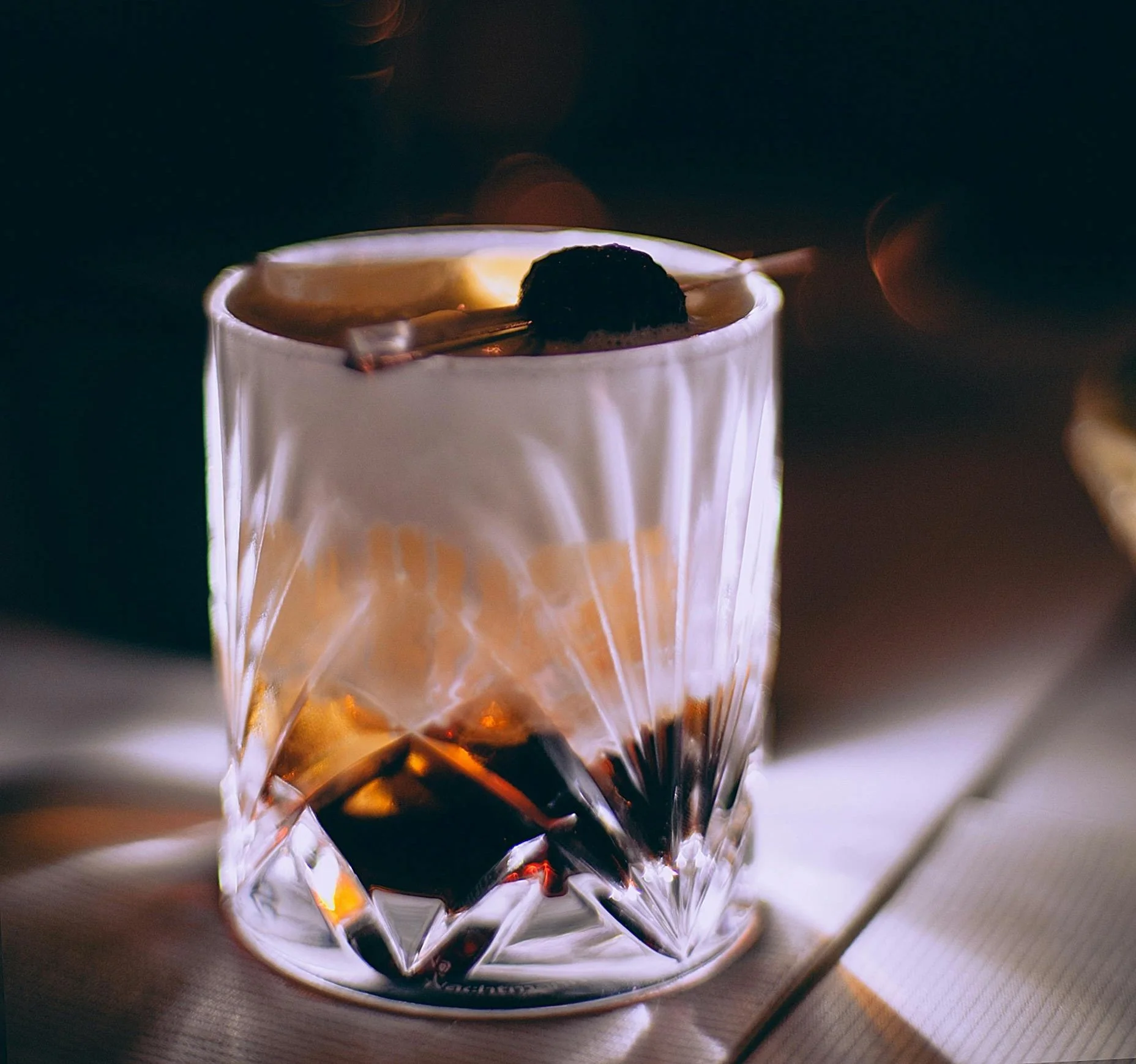 Close-up of a glass of whiskey with a large ice cube and a cherry on top, on a wooden surface.